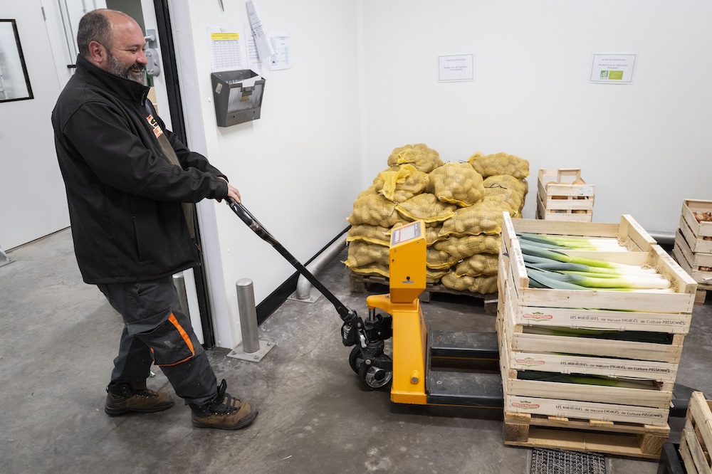 Un homme déplace des cagettes de légumes avec un transpalette dans un entrepôt.