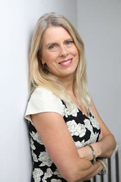 Femme blonde souriante appuyée contre un mur blanc, regardant l'objectif.