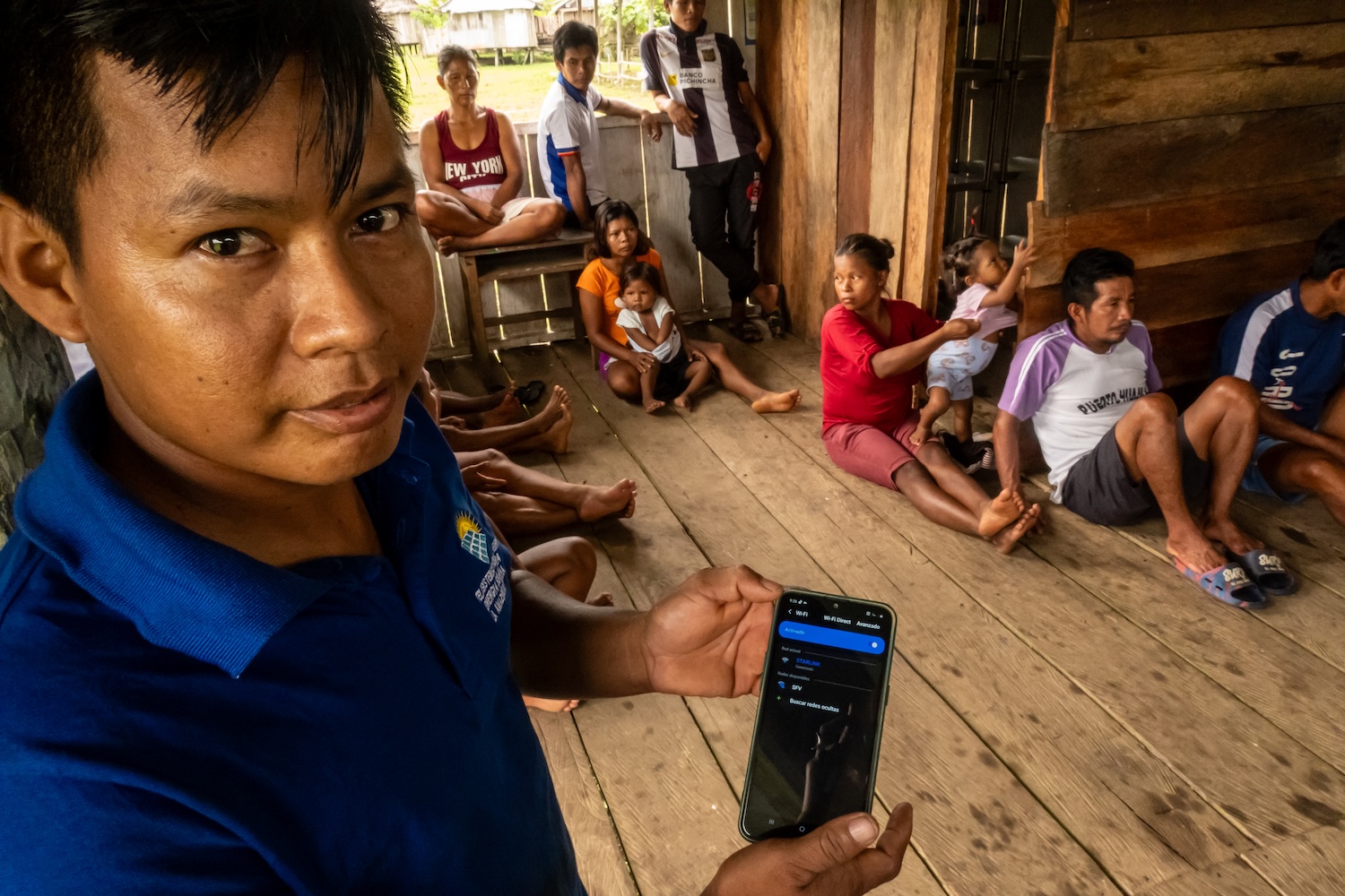 Un homme montre son téléphone dans une pièce en bois avec plusieurs personnes assises au sol.