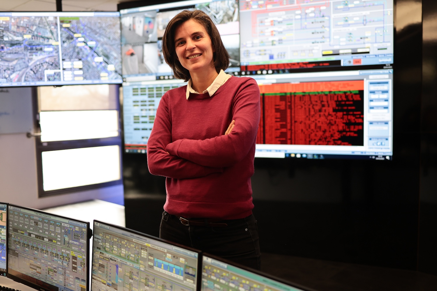 Femme souriante debout devant plusieurs écrans de contrôle dans une salle de surveillance.
