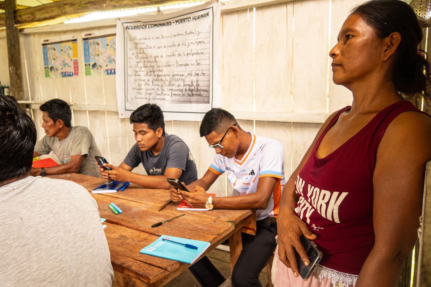Des personnes assises à une table en bois, certaines utilisent leur téléphone, une femme debout.