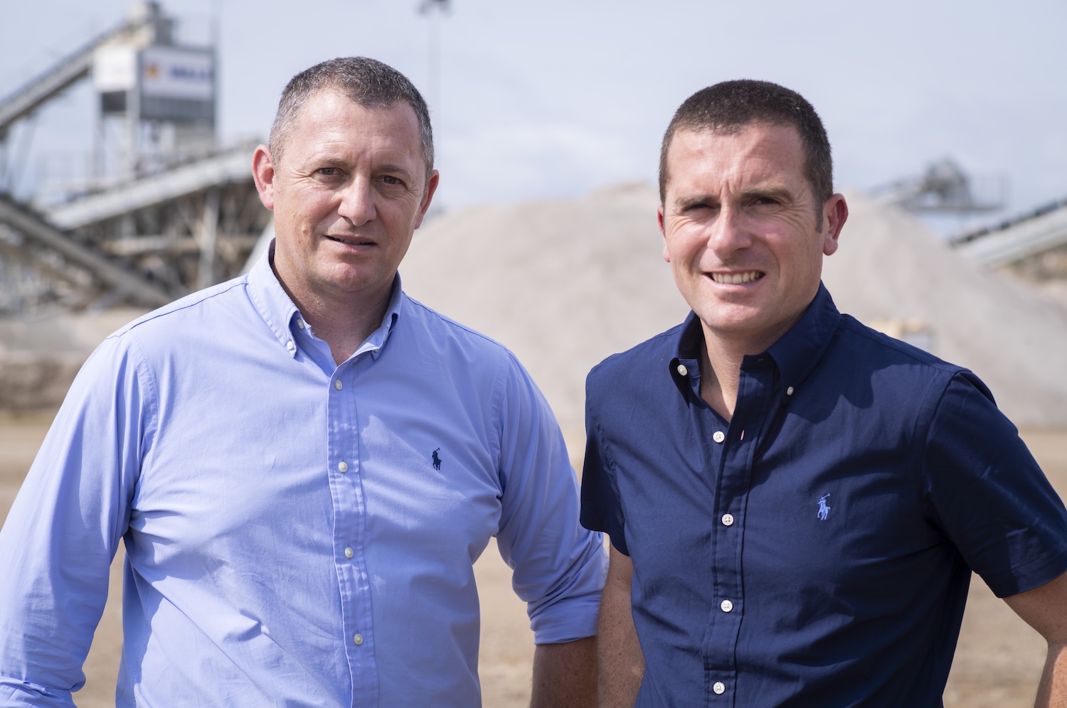 Deux hommes souriants posent devant un site industriel en plein air, sous un ciel dégagé.