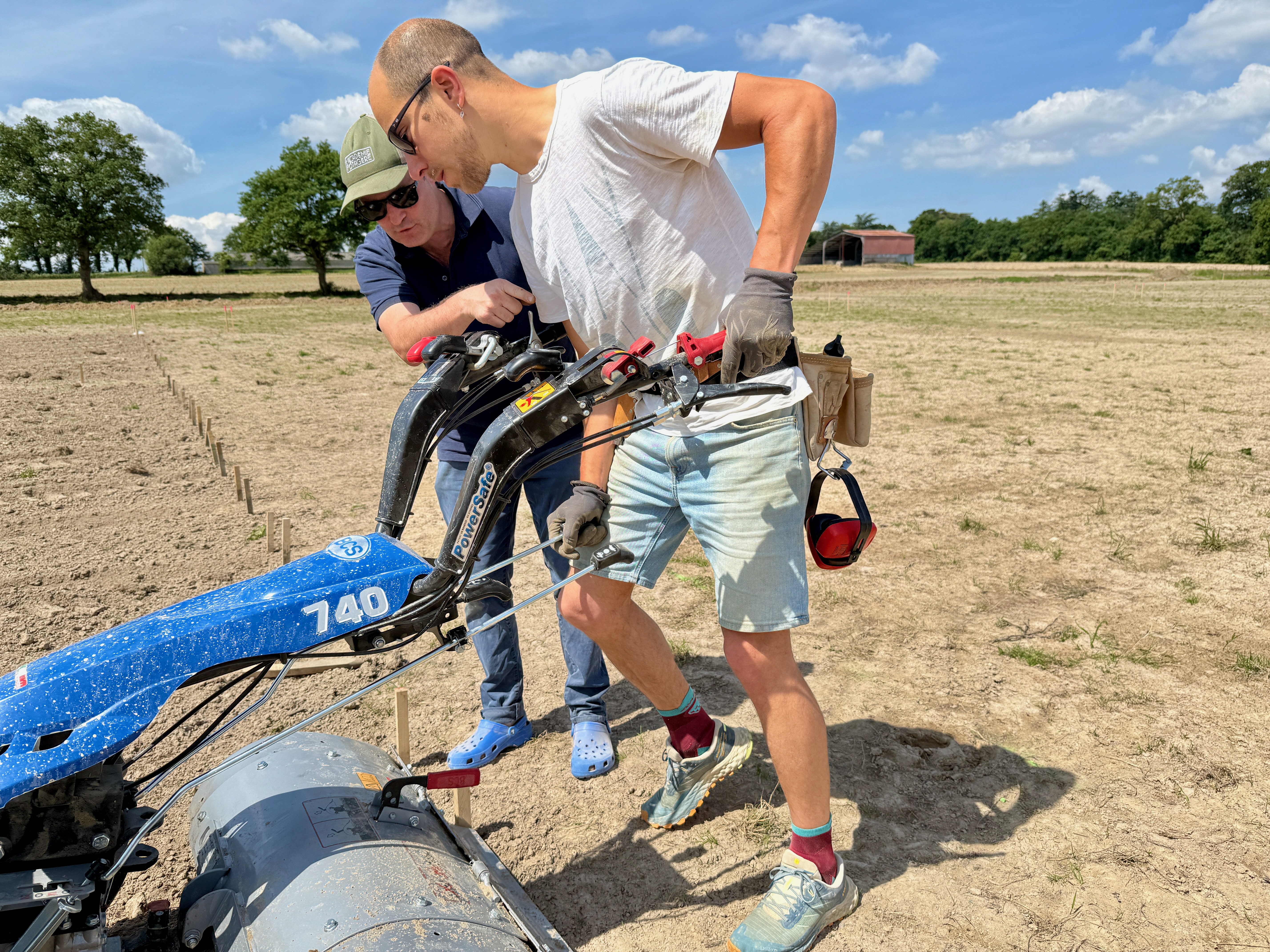 Deux hommes manipulent une machine agricole bleue dans un champ sous un ciel ensoleillé.