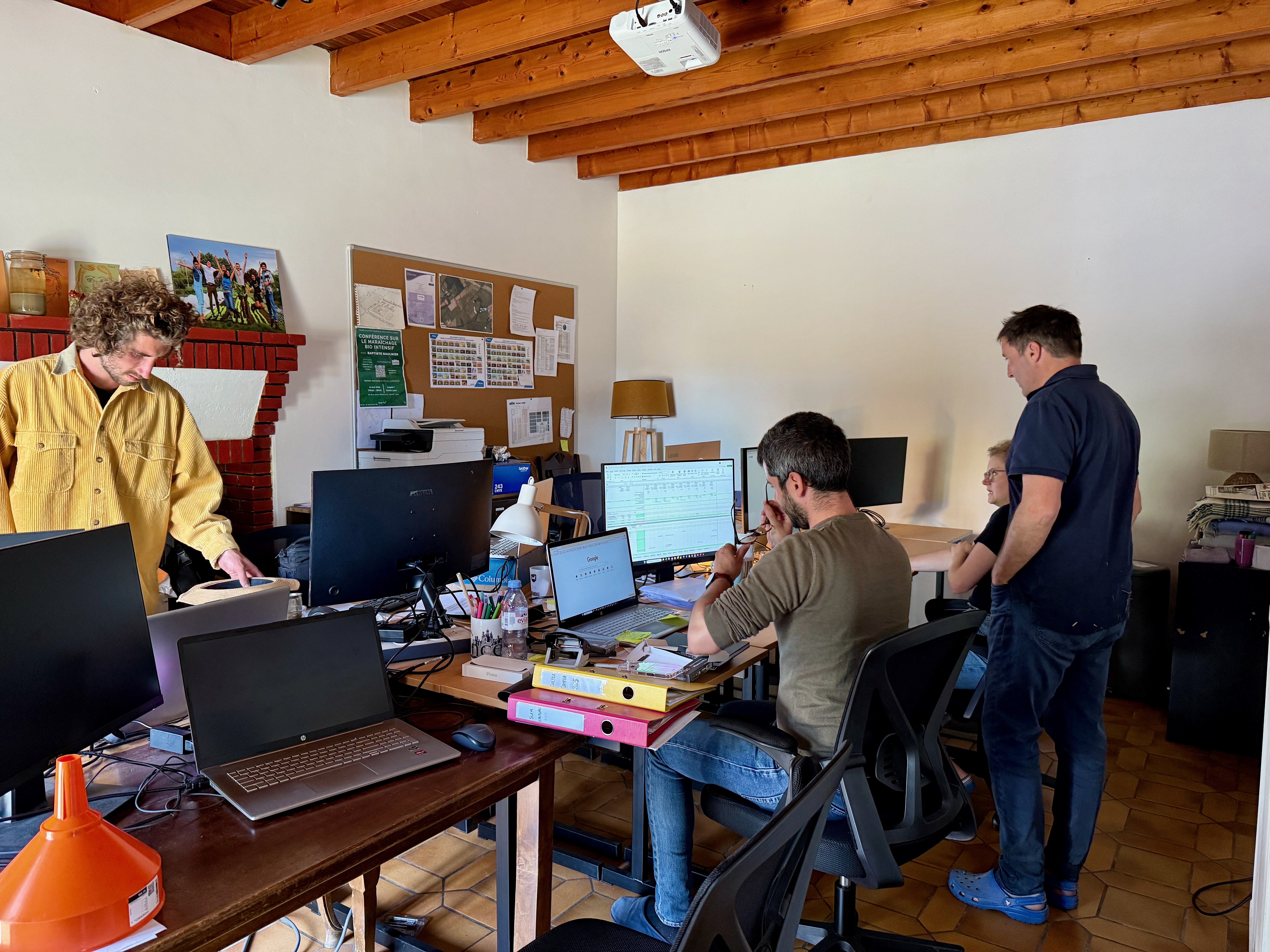 Quatre personnes travaillent dans un bureau avec plusieurs ordinateurs et des documents sur les tables.