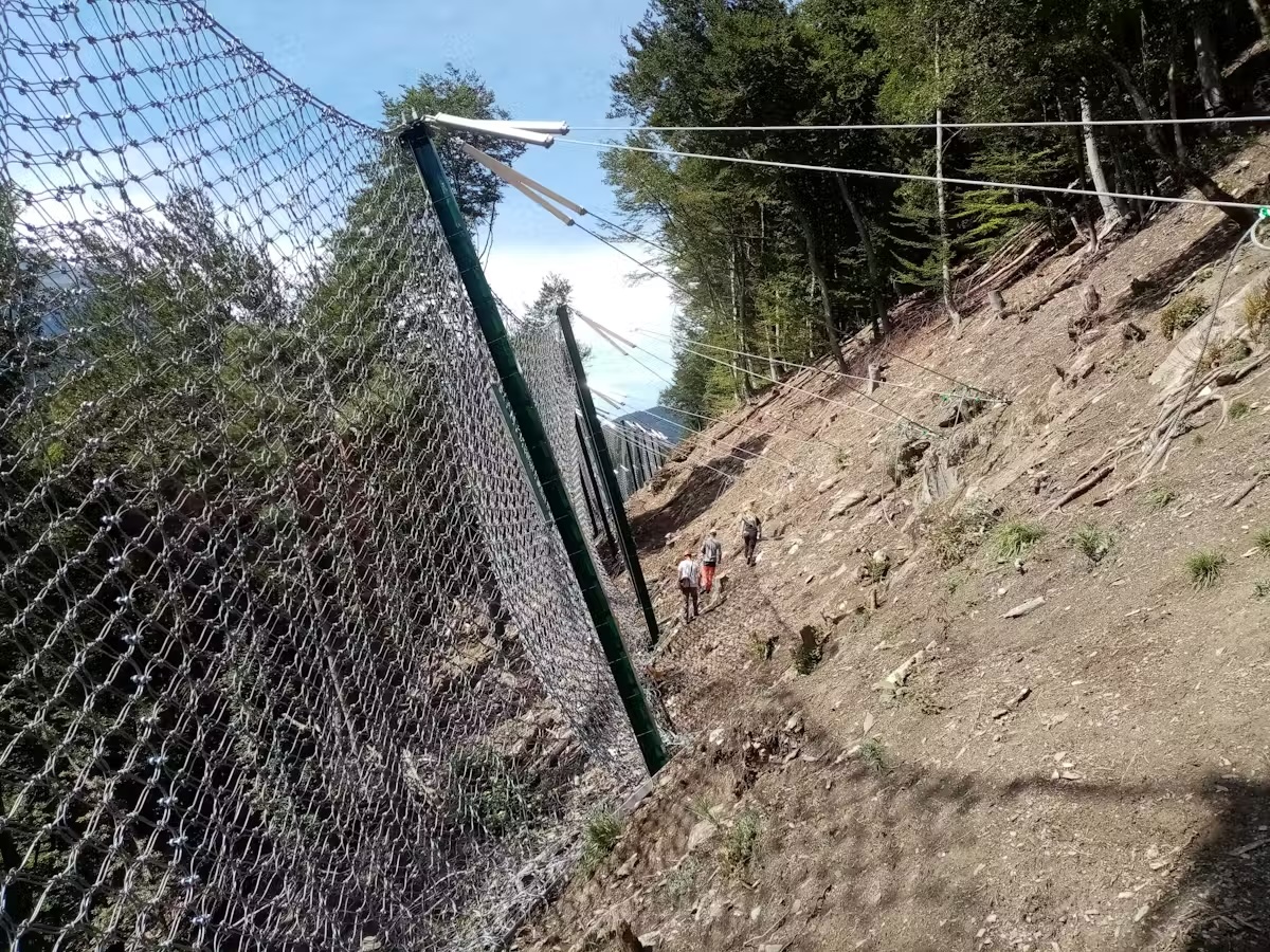Clôture de protection en montagne avec des randonneurs sur un sentier en pente boisée.