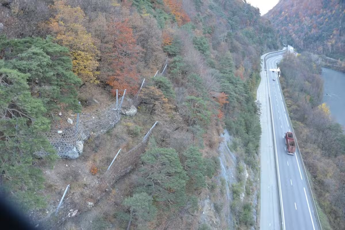 Route longeant une falaise boisée avec un camion et des filets de protection contre les éboulements.