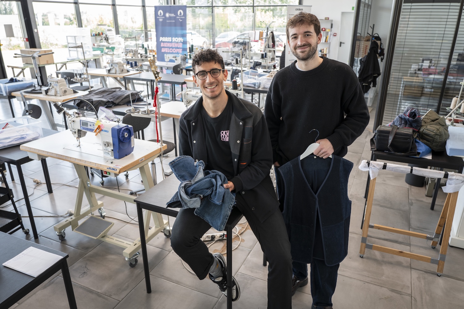 Deux hommes souriants tiennent des vêtements dans un atelier de couture moderne et lumineux.