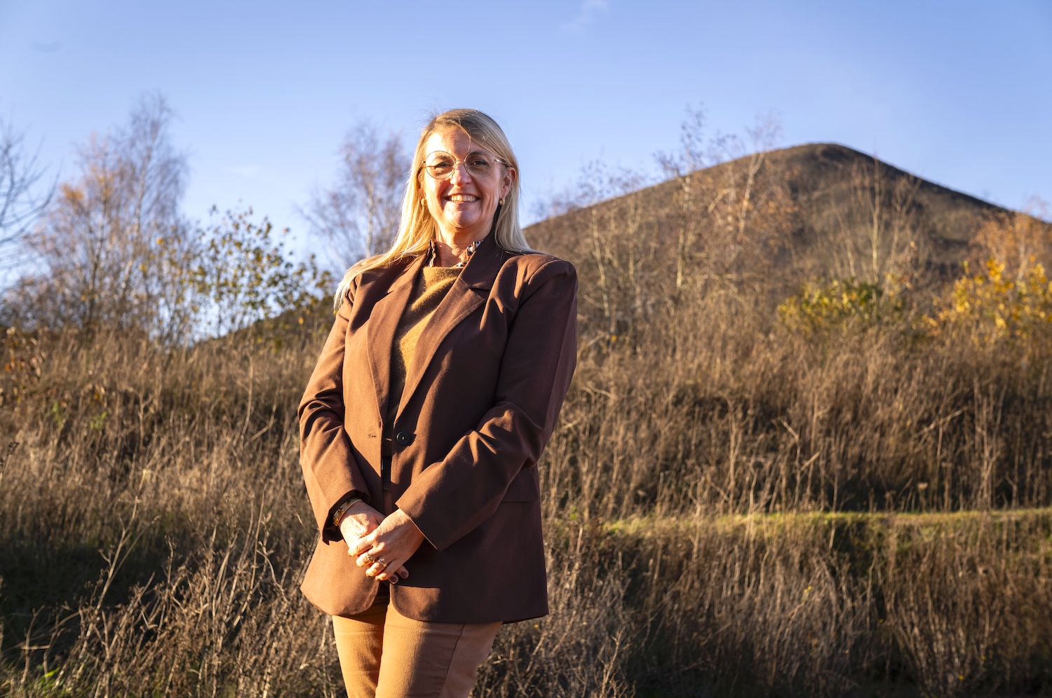 Femme souriante en costume marron devant une colline sous un ciel bleu clair