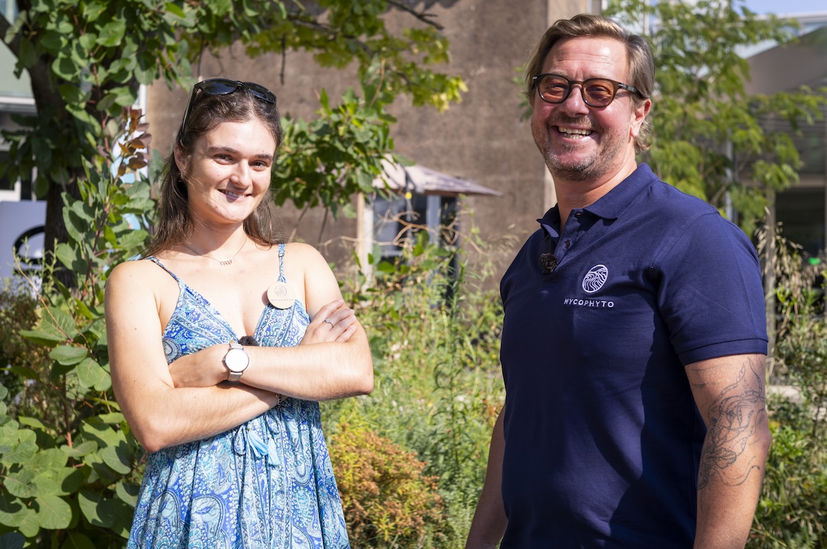 Deux personnes souriantes posent dehors, sous le soleil, devant un jardin verdoyant.