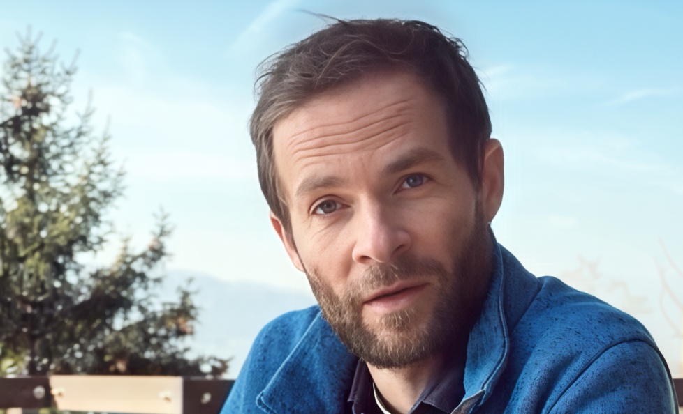 Homme barbu en veste bleue assis dehors avec un arbre et ciel bleu en arrière-plan