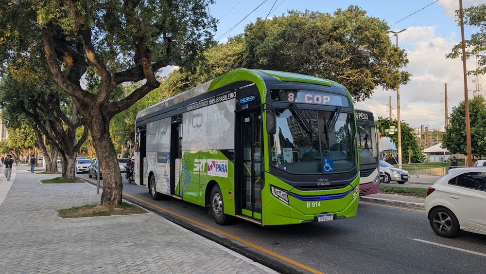 Bus vert électrique circulant sur une avenue bordée d'arbres en pleine journée.