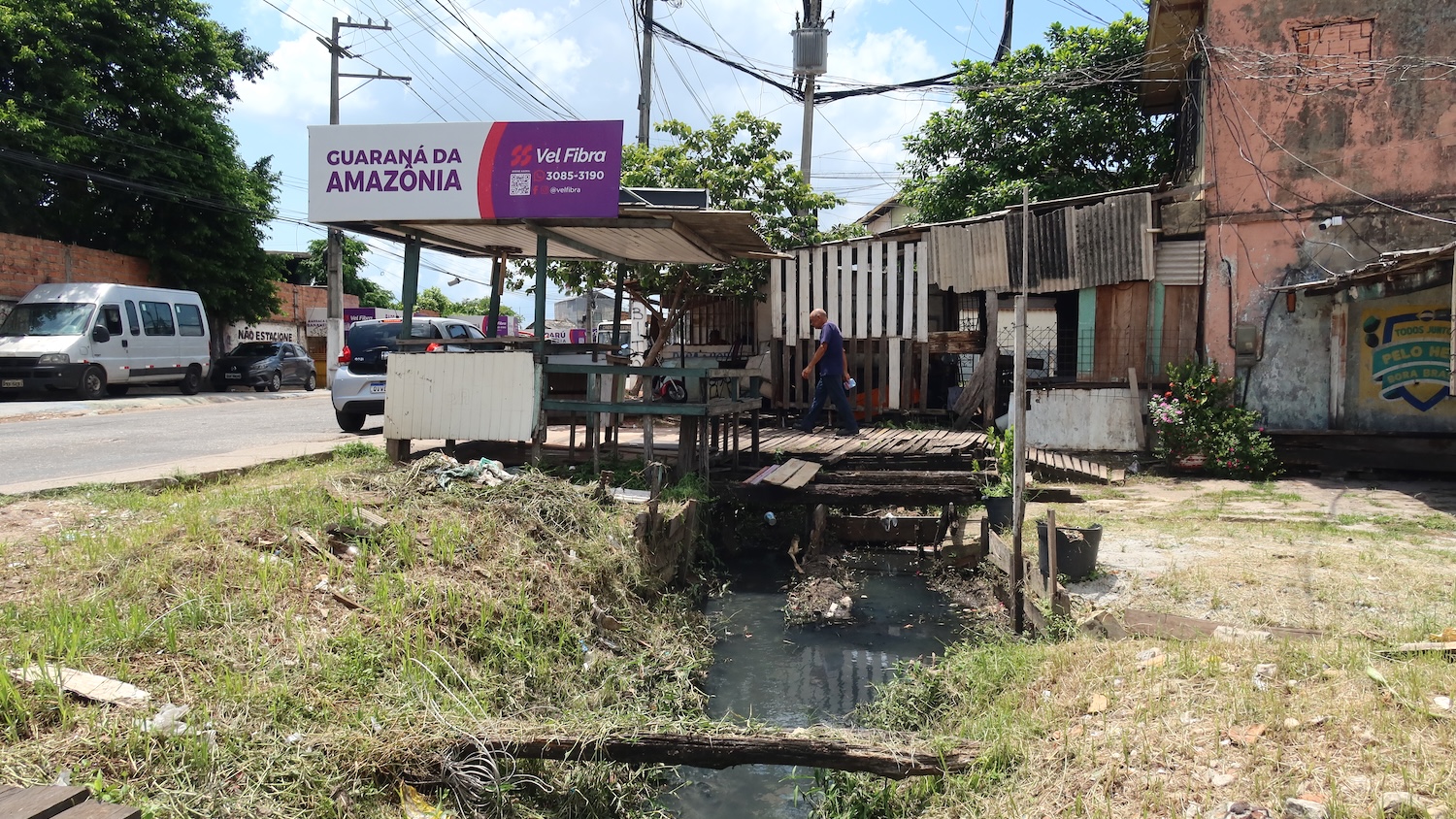 Échoppe en bois au bord d’un égout à ciel ouvert, près d’une route fréquentée.
