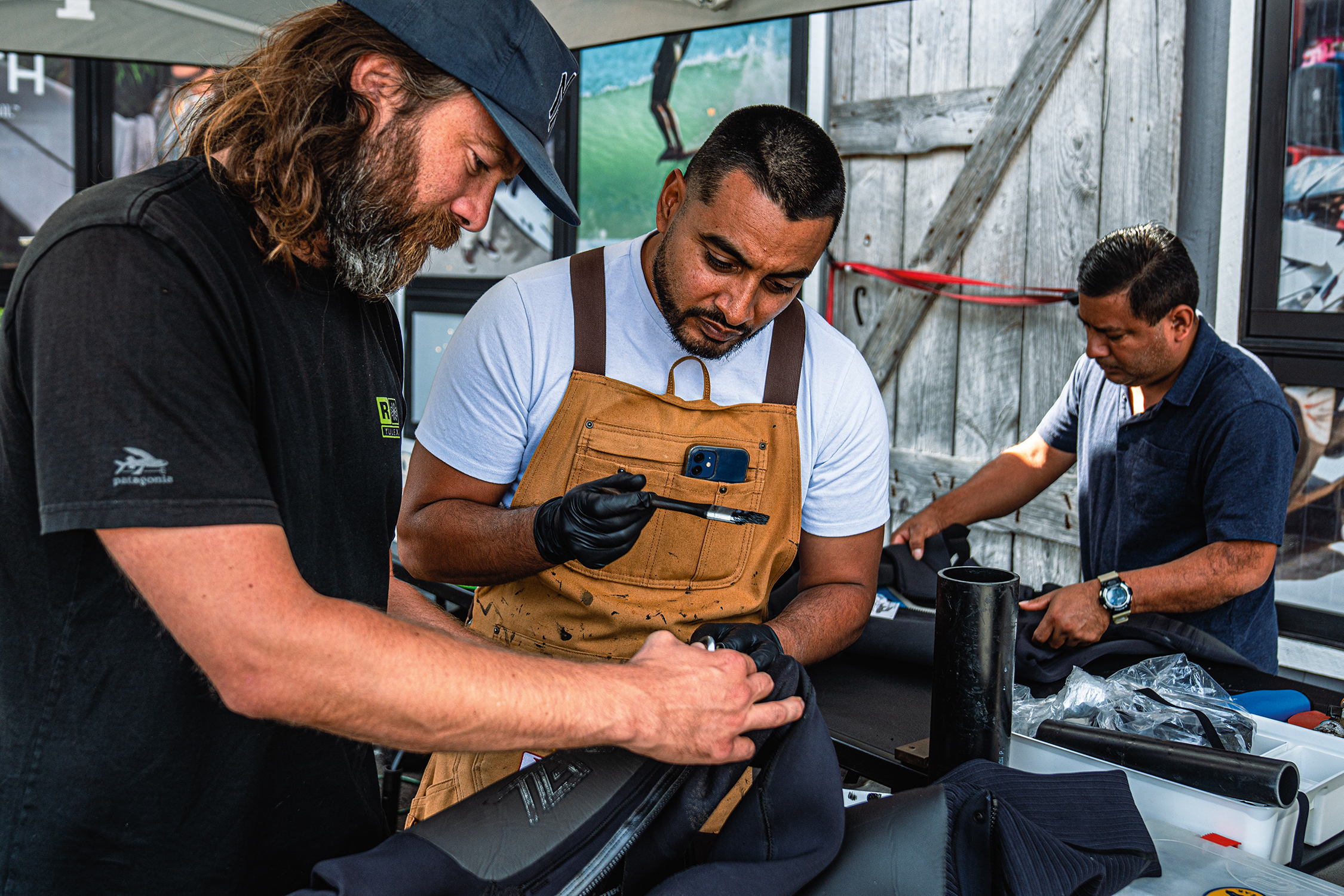 Trois hommes concentrés réparent ou assemblent des pièces mécaniques sur un établi en extérieur.