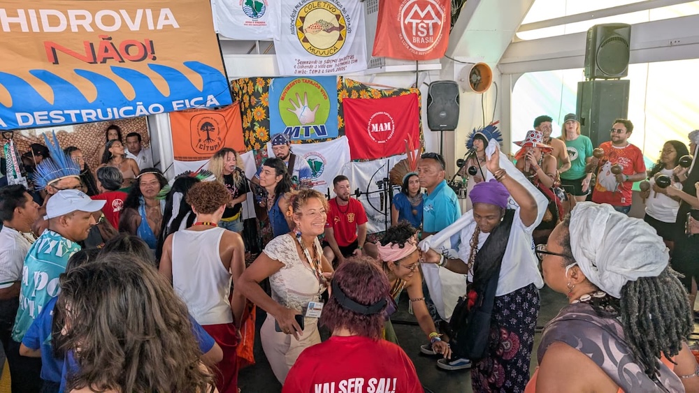 Groupe de personnes dansant joyeusement lors d’un rassemblement avec des banderoles militantes en arrière-plan.