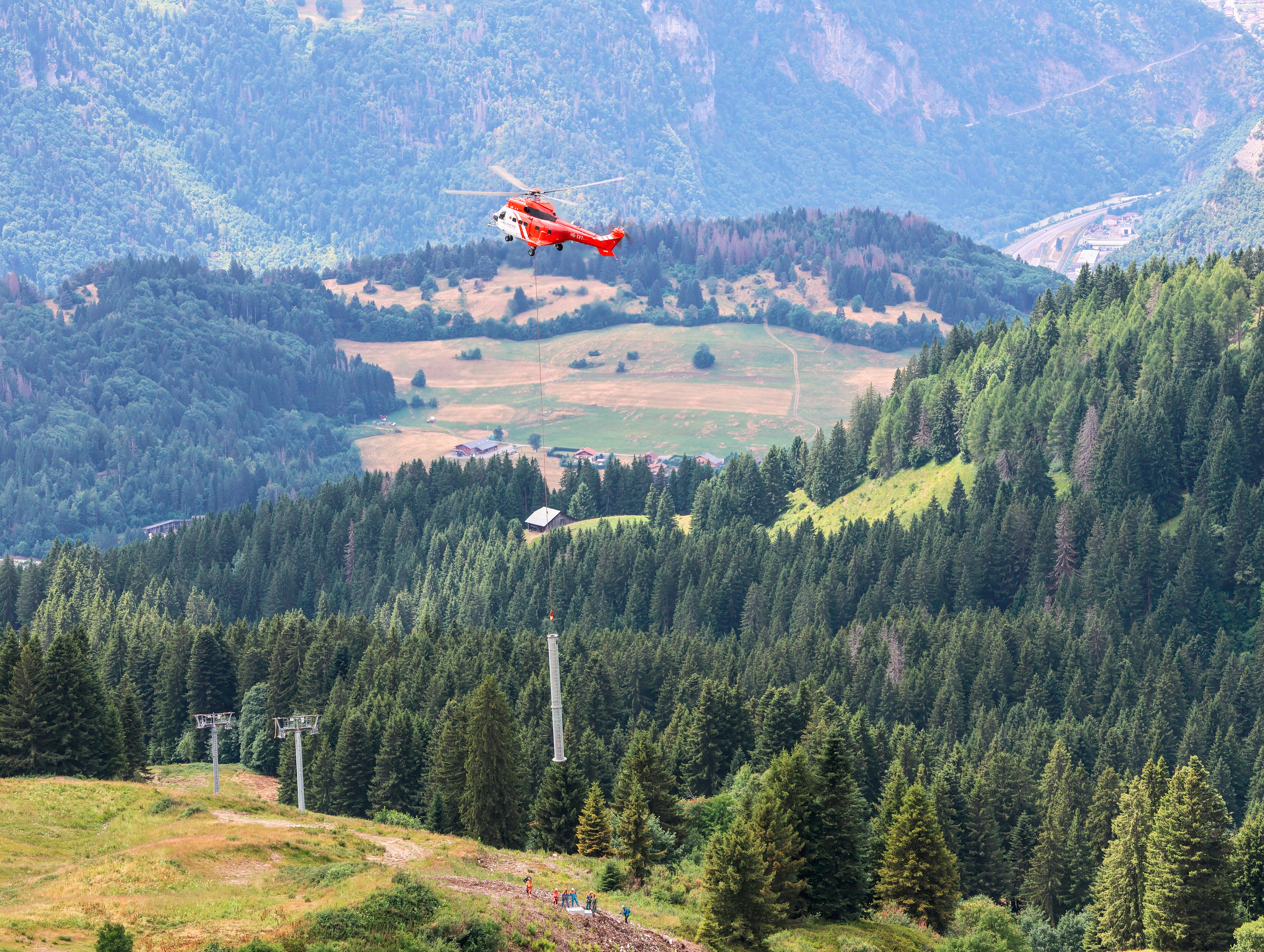 Hélicoptère rouge survolant une vallée montagneuse verdoyante avec forêts et prairies.