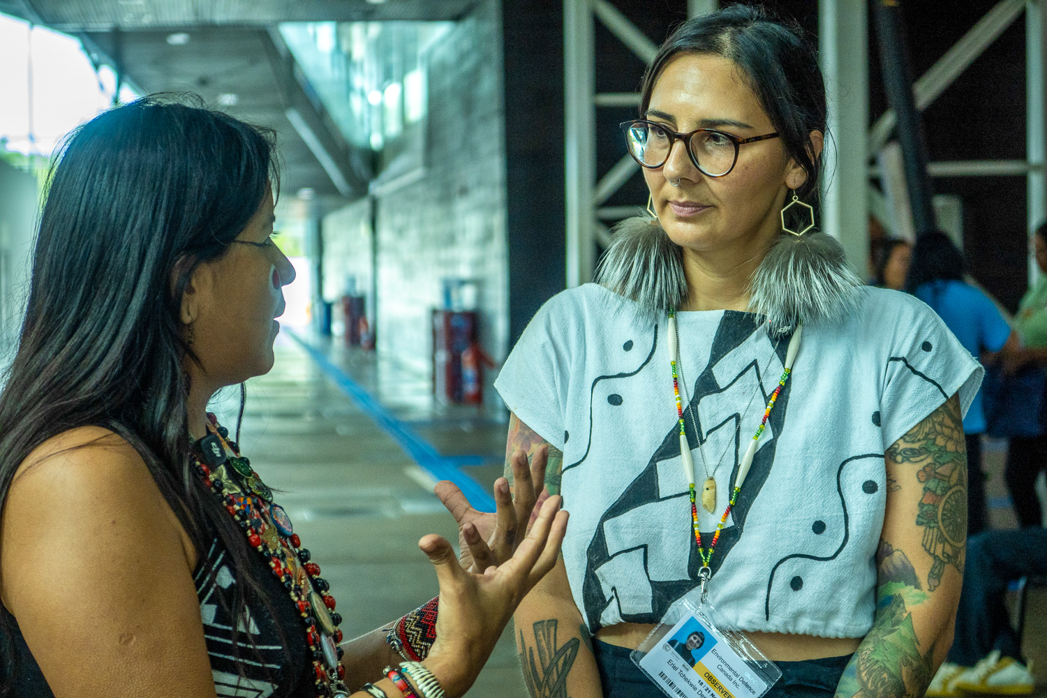 Deux femmes discutent dans un couloir moderne, vêtues de tenues traditionnelles et colorées.