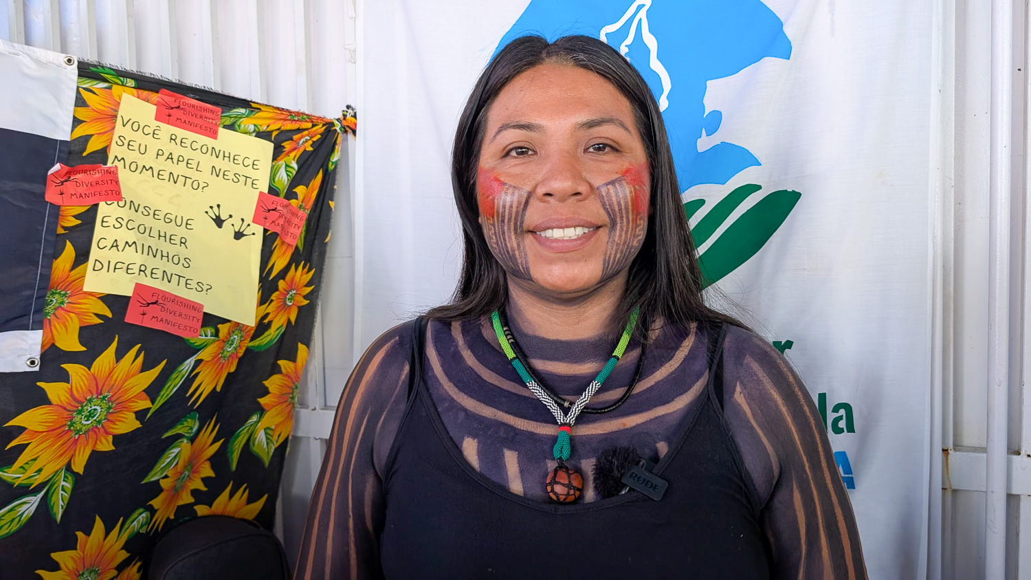 Femme souriante avec peintures traditionnelles, devant une affiche colorée et un drapeau blanc.