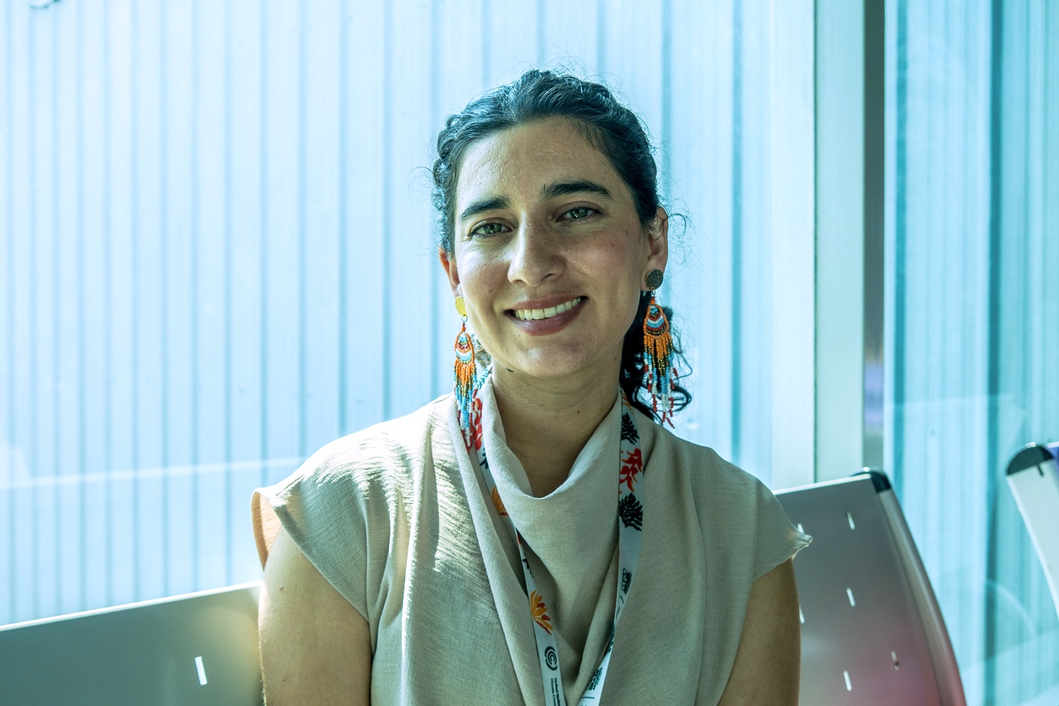 Femme souriante assise près d'une fenêtre, portant des boucles d'oreilles colorées et un foulard.