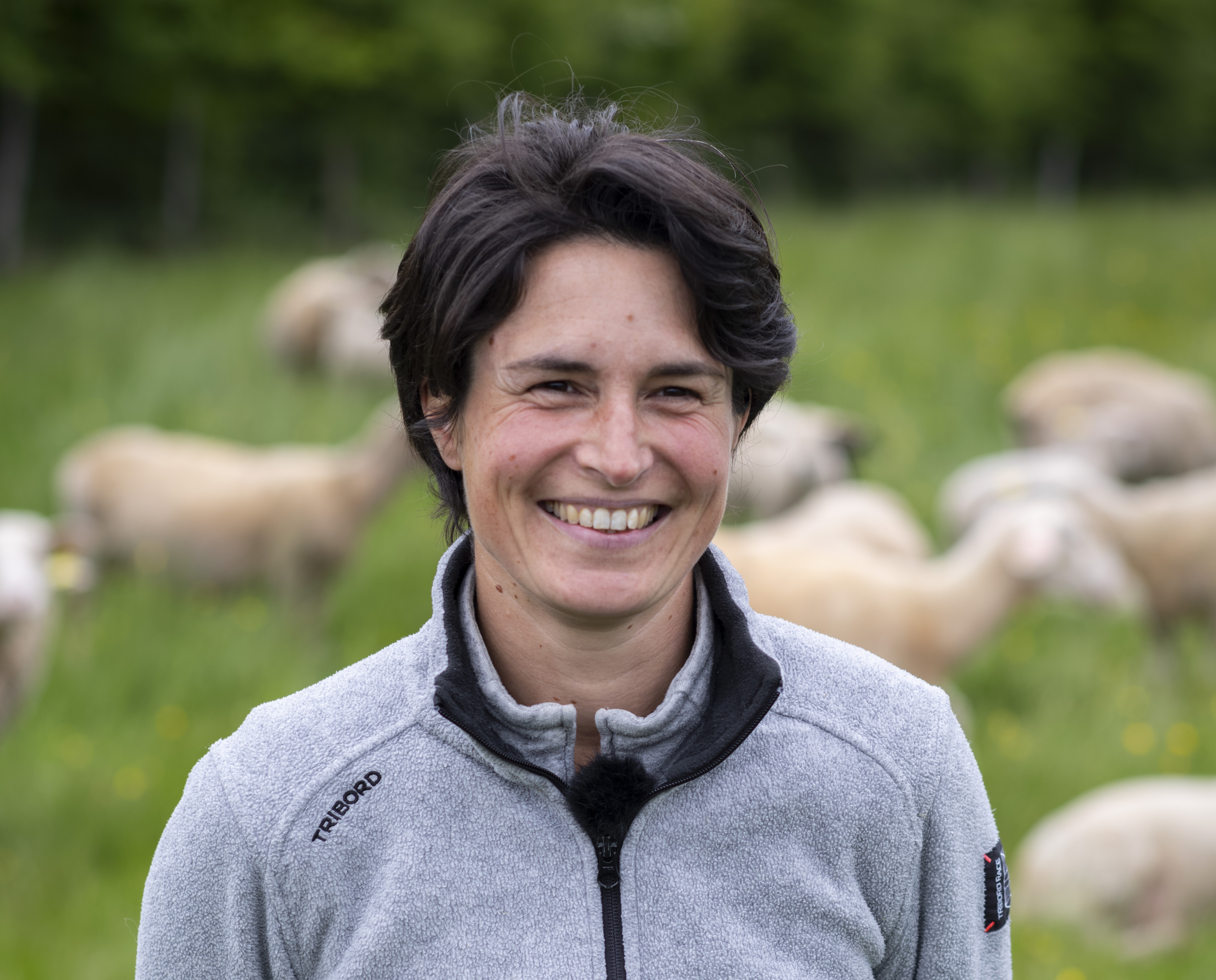 Femme souriante, Pascaline Loquet, devant un troupeau de moutons dans un champ verdoyant.