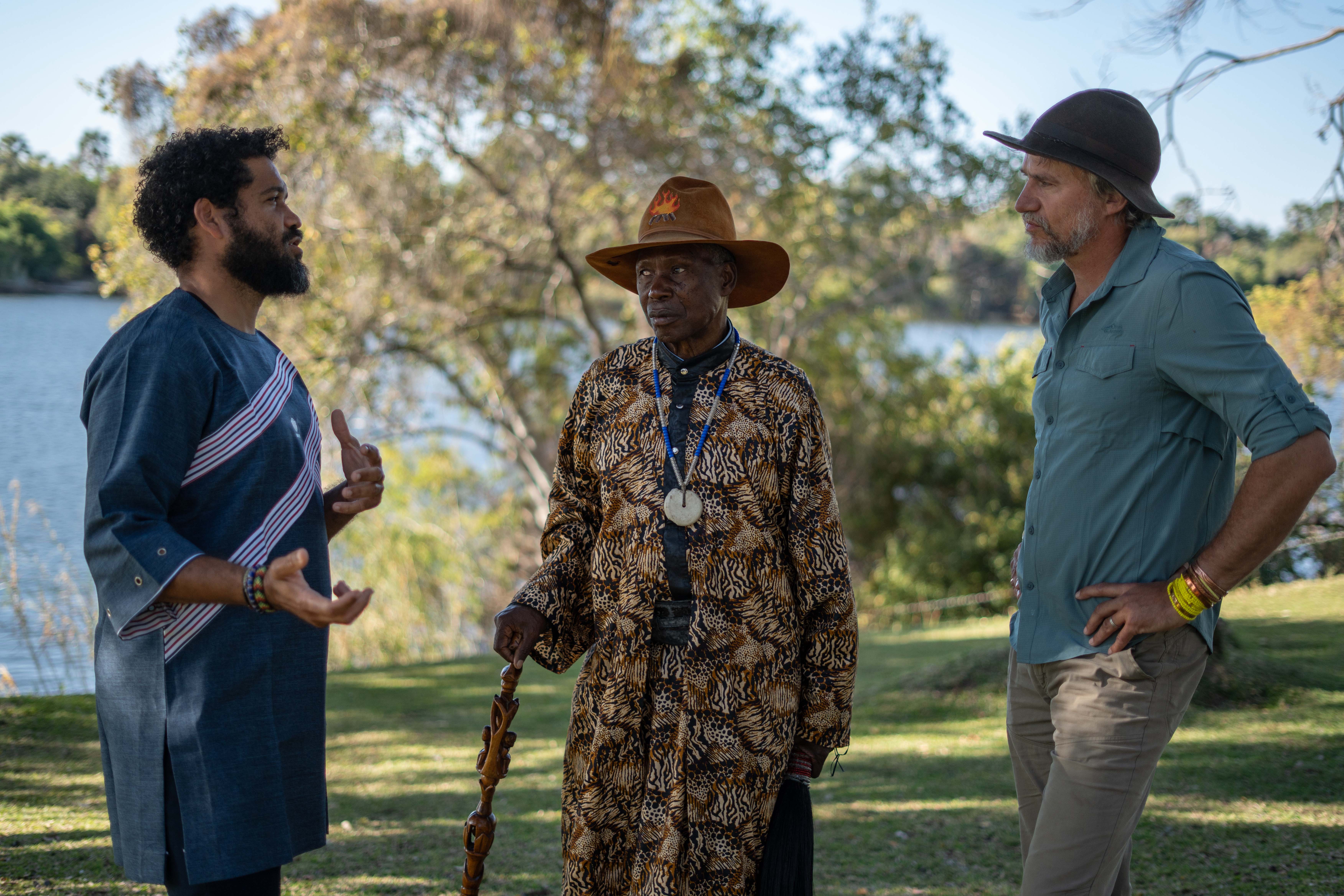 Trois hommes discutent en plein air près d’un lac, entourés d’arbres et de verdure.