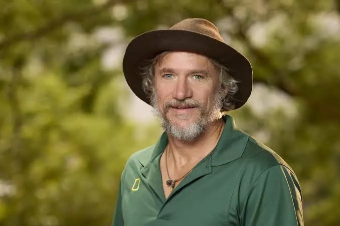 Homme barbu portant un chapeau marron et une chemise verte, en extérieur verdoyant.
