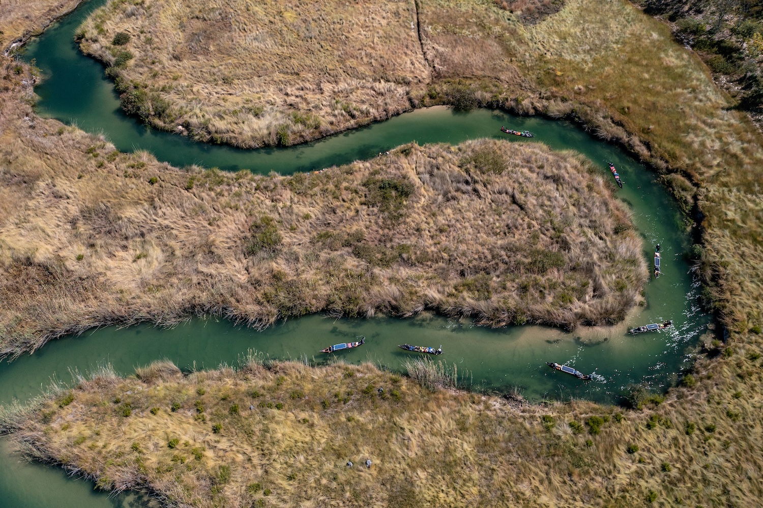 Vue aérienne d'une rivière sinueuse entourée de végétation sèche avec des canoës sur l'eau.