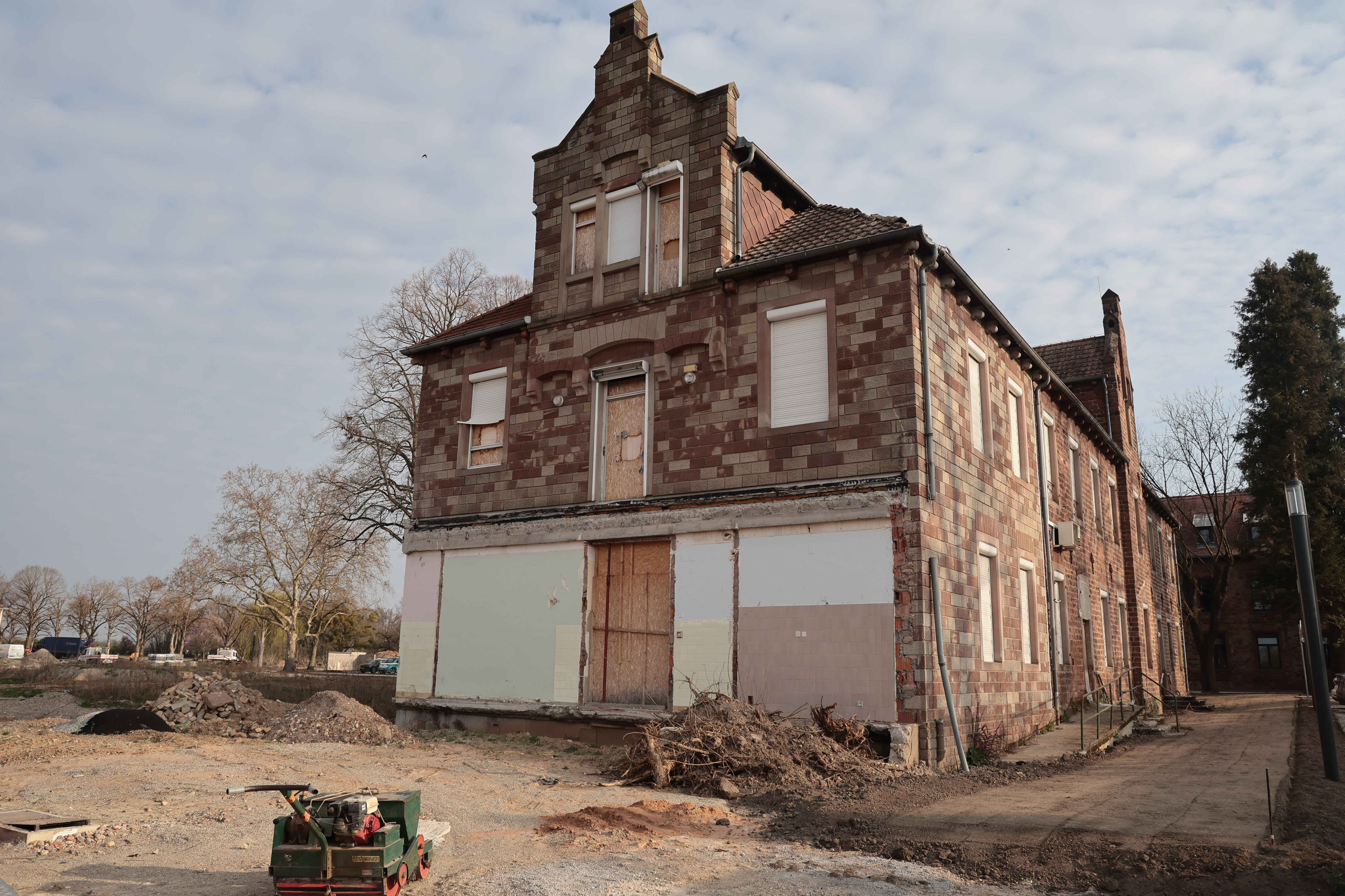 Bâtiment en pierre partiellement muré, entouré de terre et matériel de chantier, ciel partiellement nuageux.