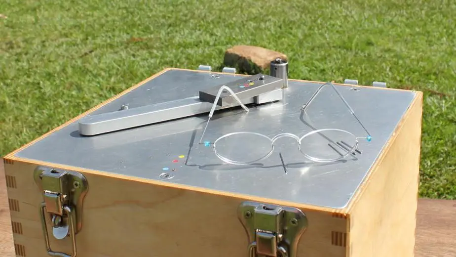 Boîte en bois avec un pantographe métallique posé sur le dessus, en extérieur sur l'herbe.