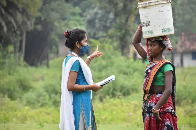 Une femme interroge une villageoise portant une boîte sur la tête dans un cadre rural.