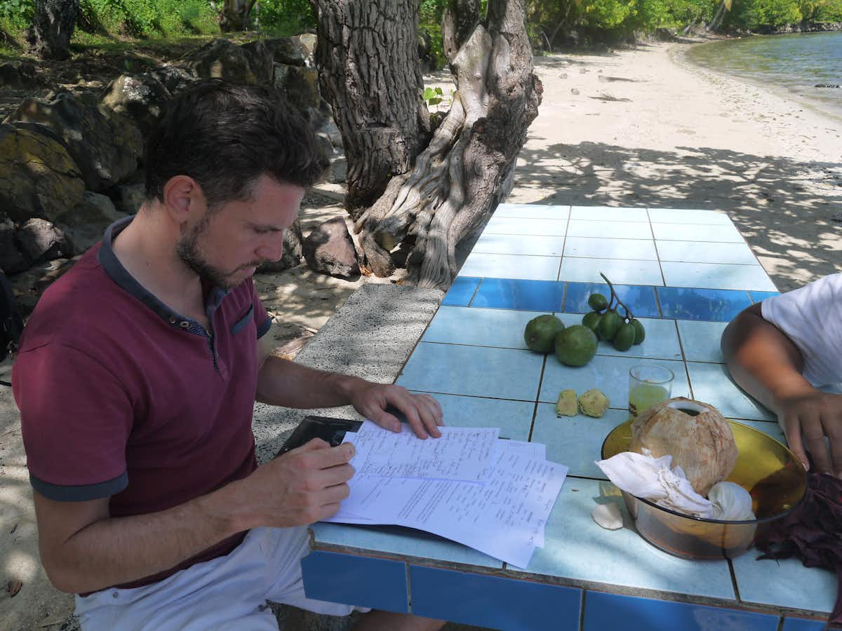 Homme écrivant sur des feuilles à une table en bord de plage, fruits tropicaux posés dessus.