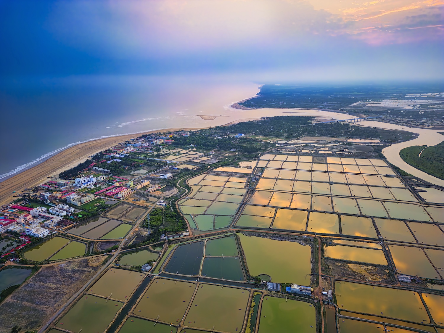 Paysage côtier avec champs rectangulaires inondés, rivière sinueuse et habitations près de la mer.