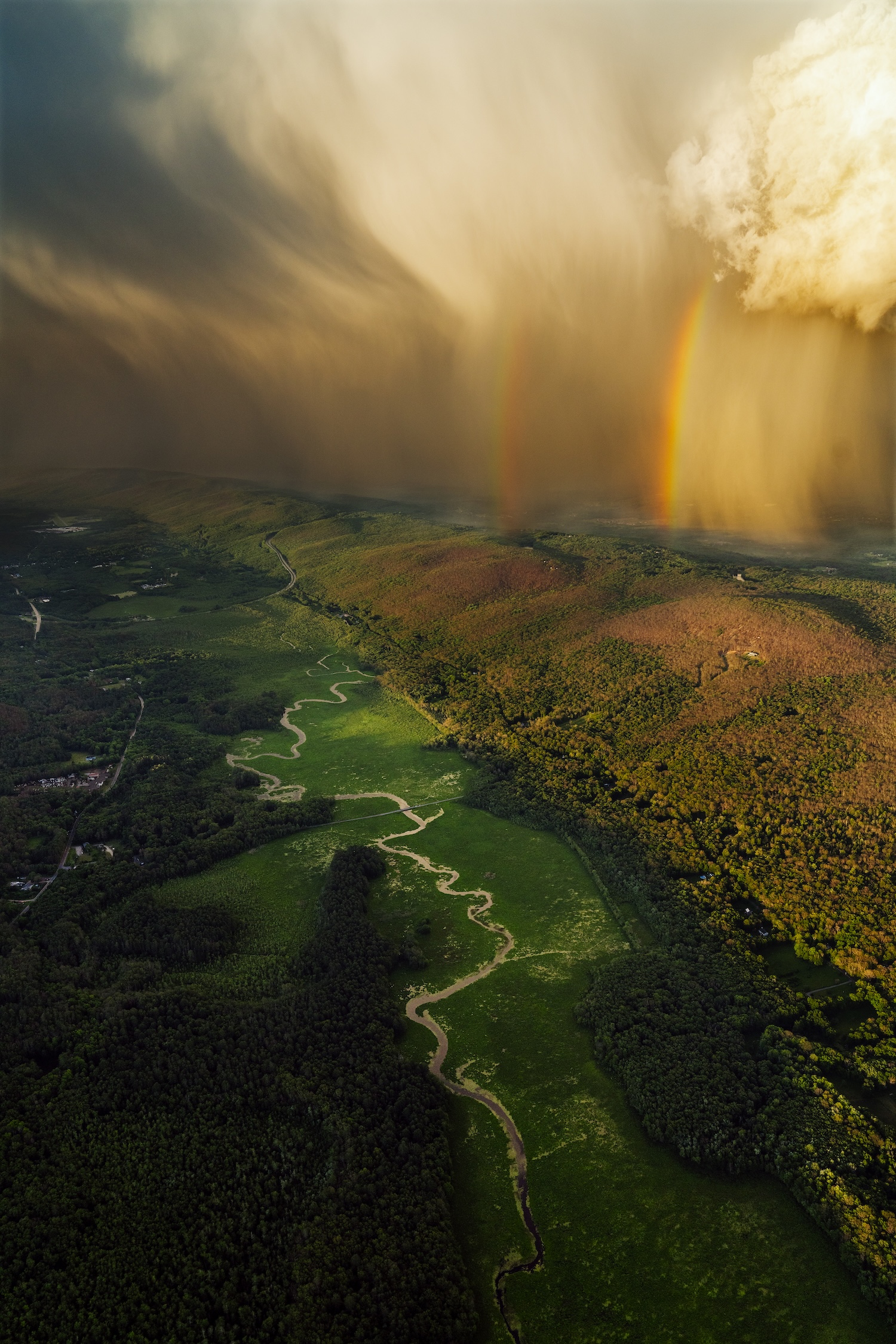 Paysage vallonné verdoyant avec rivière sinueuse et double arc-en-ciel sous un ciel orageux