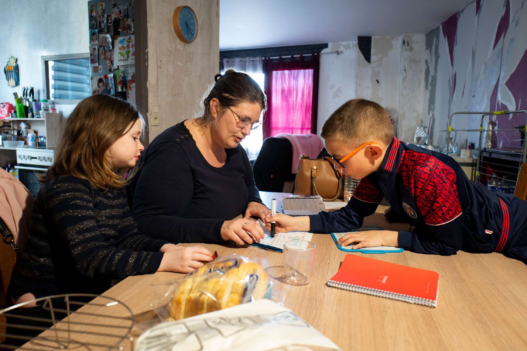 Une femme aide deux enfants à faire leurs devoirs sur une table de cuisine.