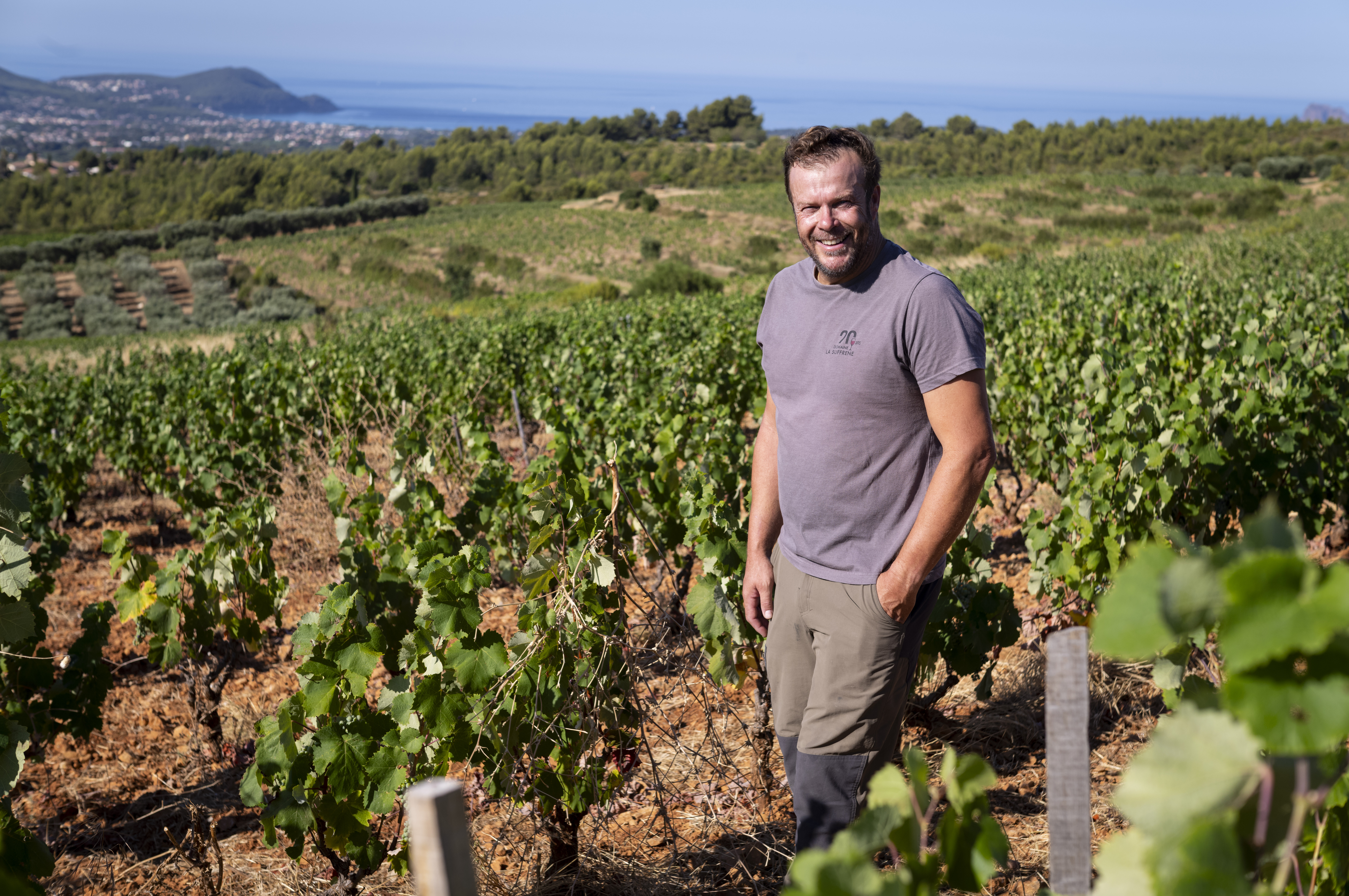Homme souriant dans un vignoble ensoleillé avec vue sur la mer et les collines.