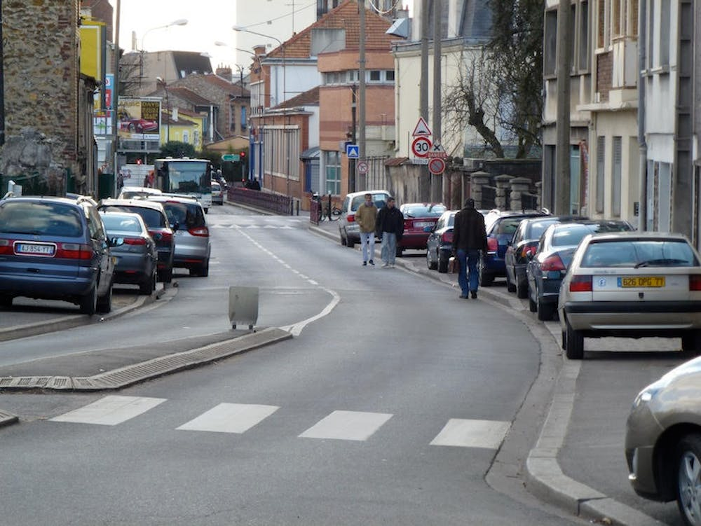 Rue étroite en ville avec voitures garées, piétons marchant sur les trottoirs et passages piétons.