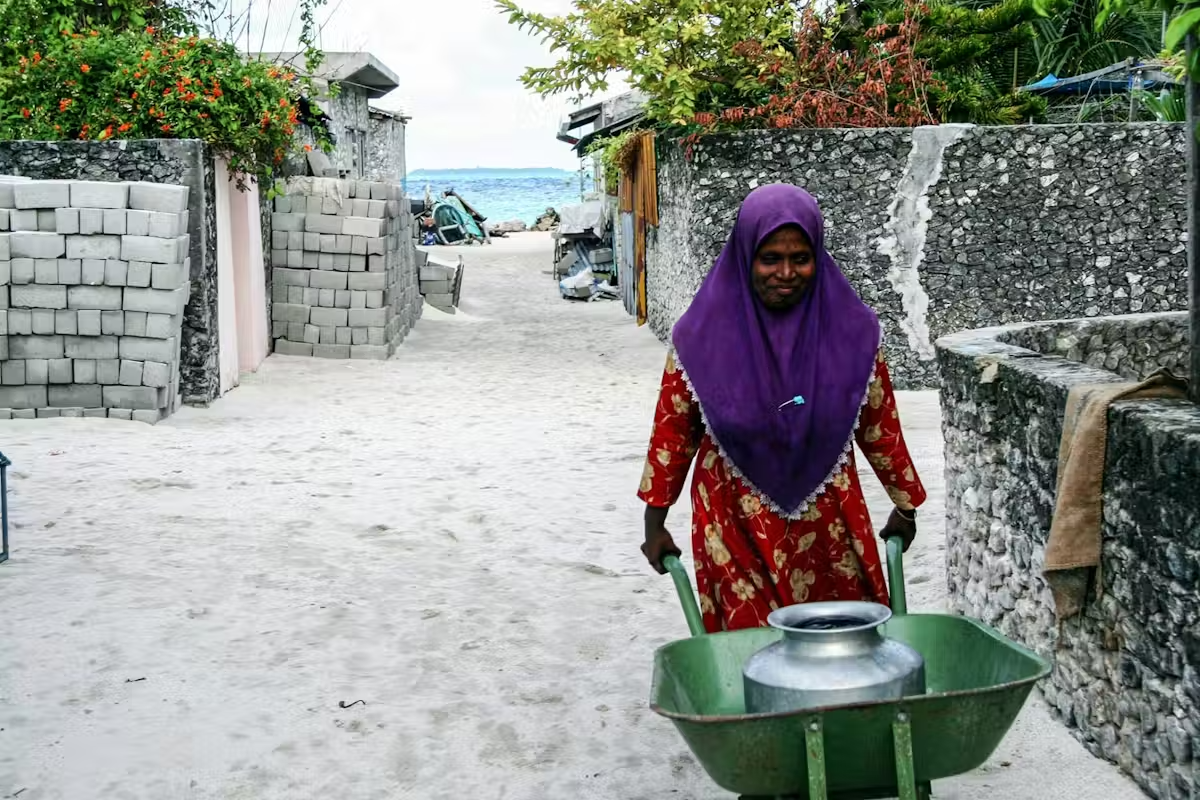 Femme en hijab violet poussant une brouette dans une ruelle sableuse près de la mer