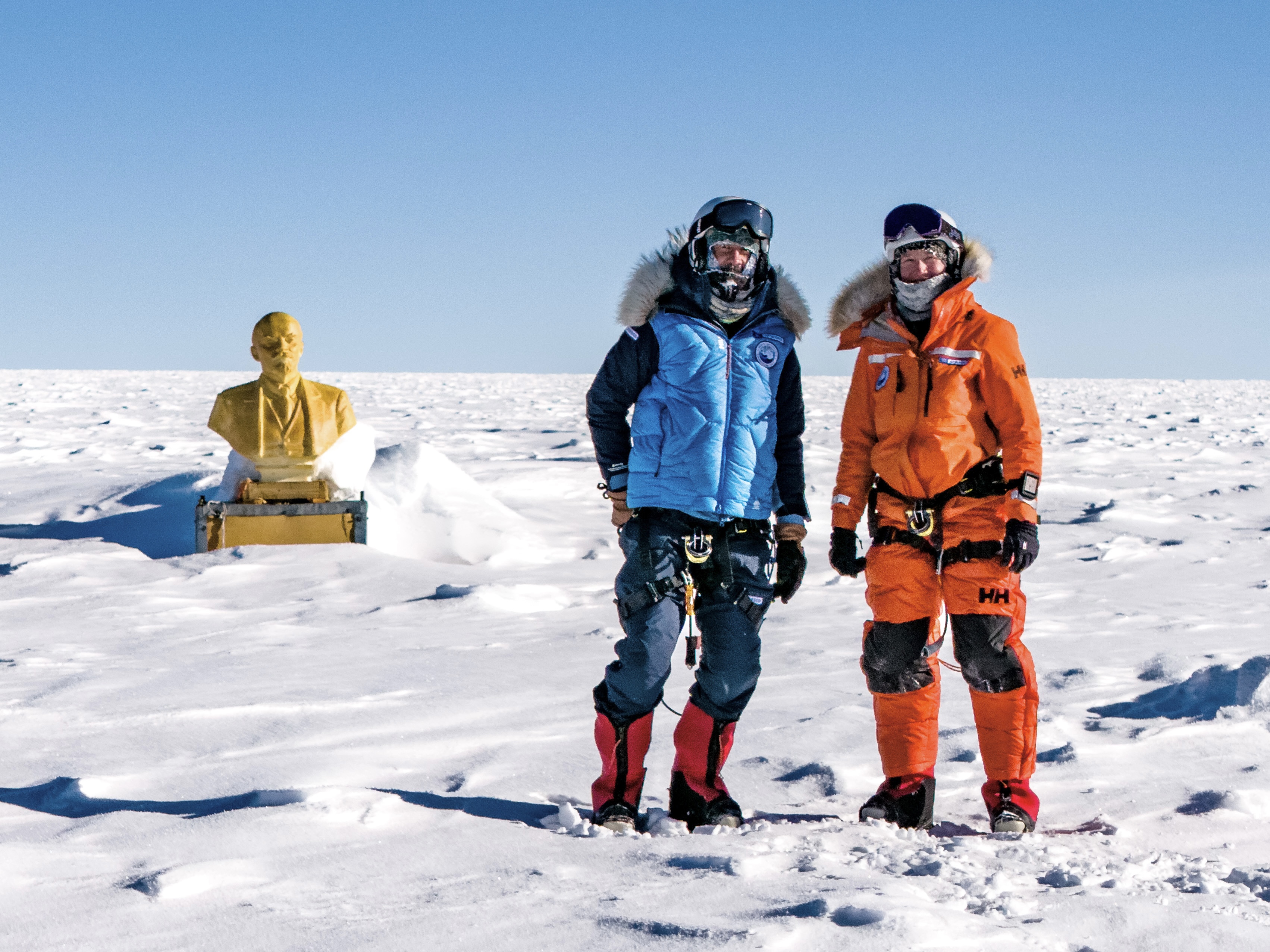 Deux explorateurs en tenue polaire posent devant une statue dorée sur la glace.