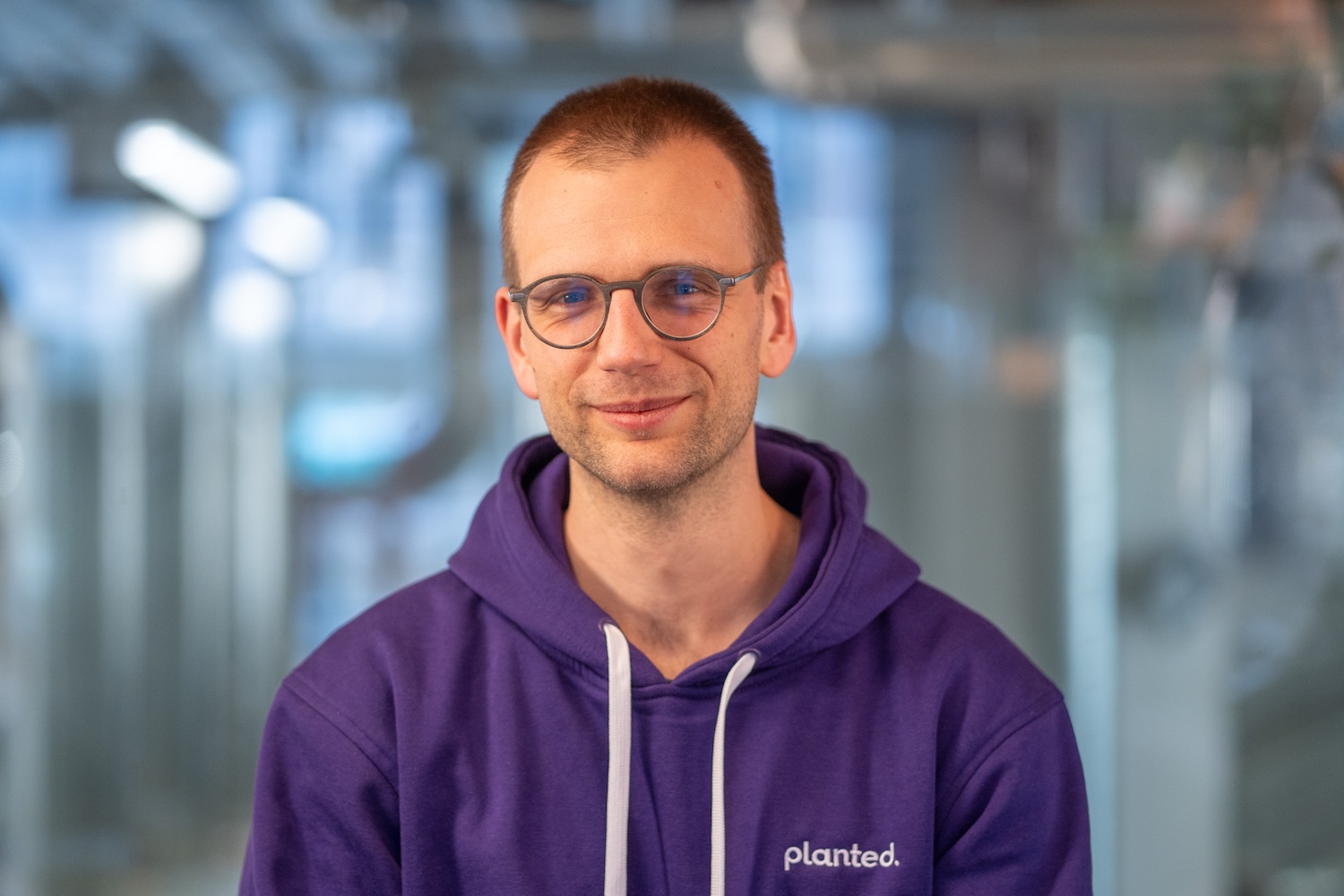 Homme souriant avec lunettes et sweat violet, en intérieur à la lumière naturelle.