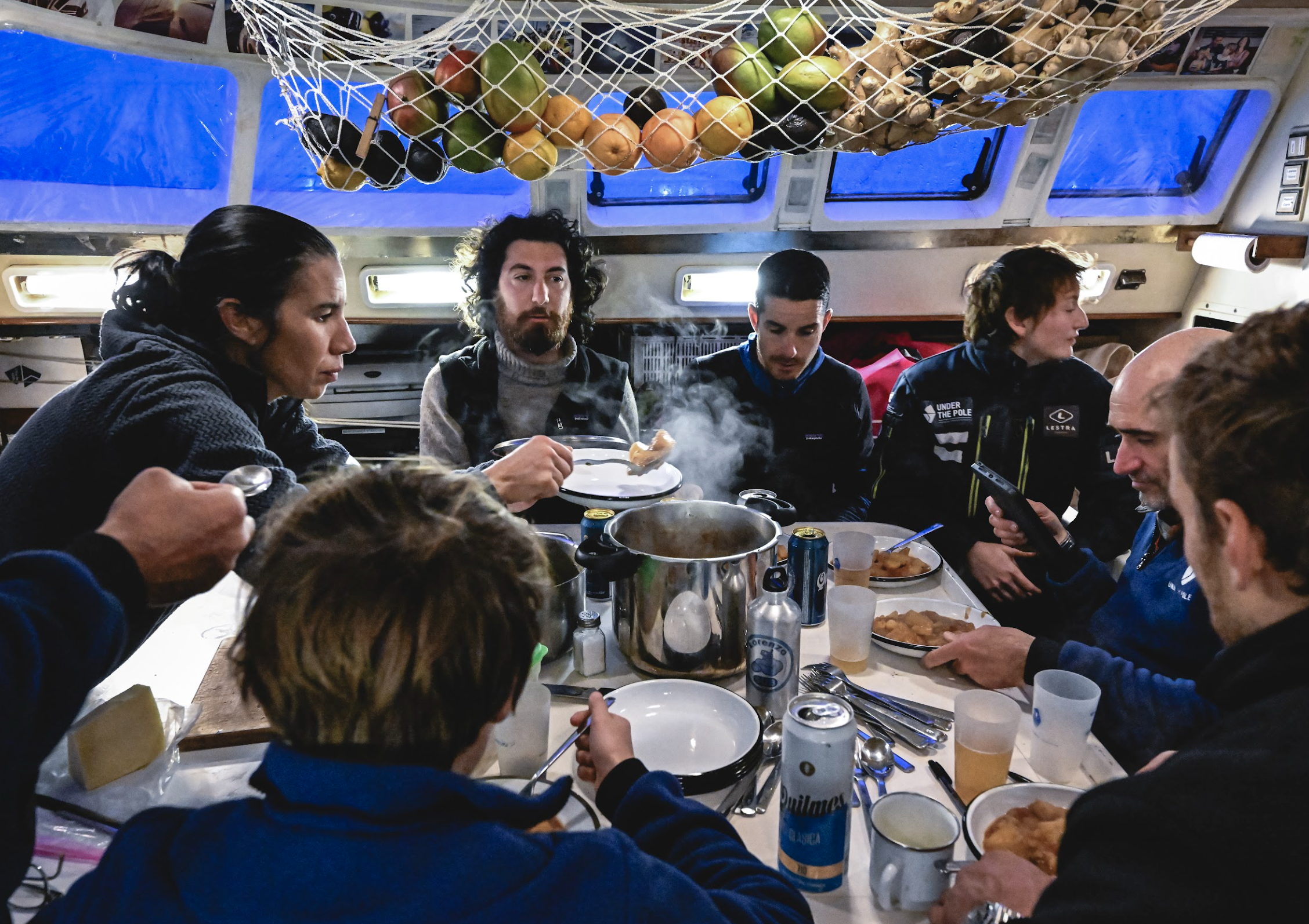 Équipage partageant un repas chaud à bord d’un bateau, fruits suspendus au plafond.