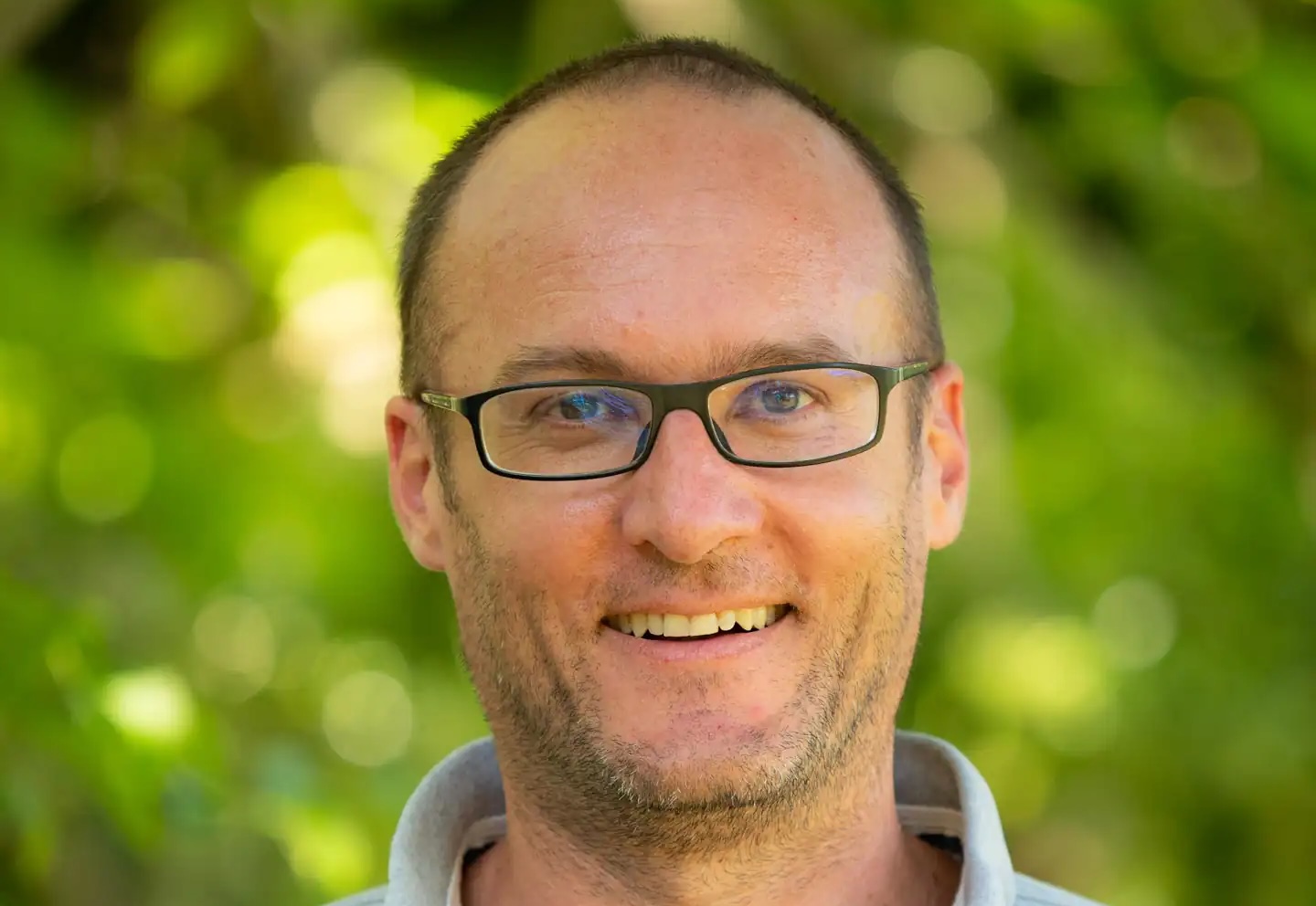 Homme souriant portant des lunettes, fond flou de verdure, chemise claire