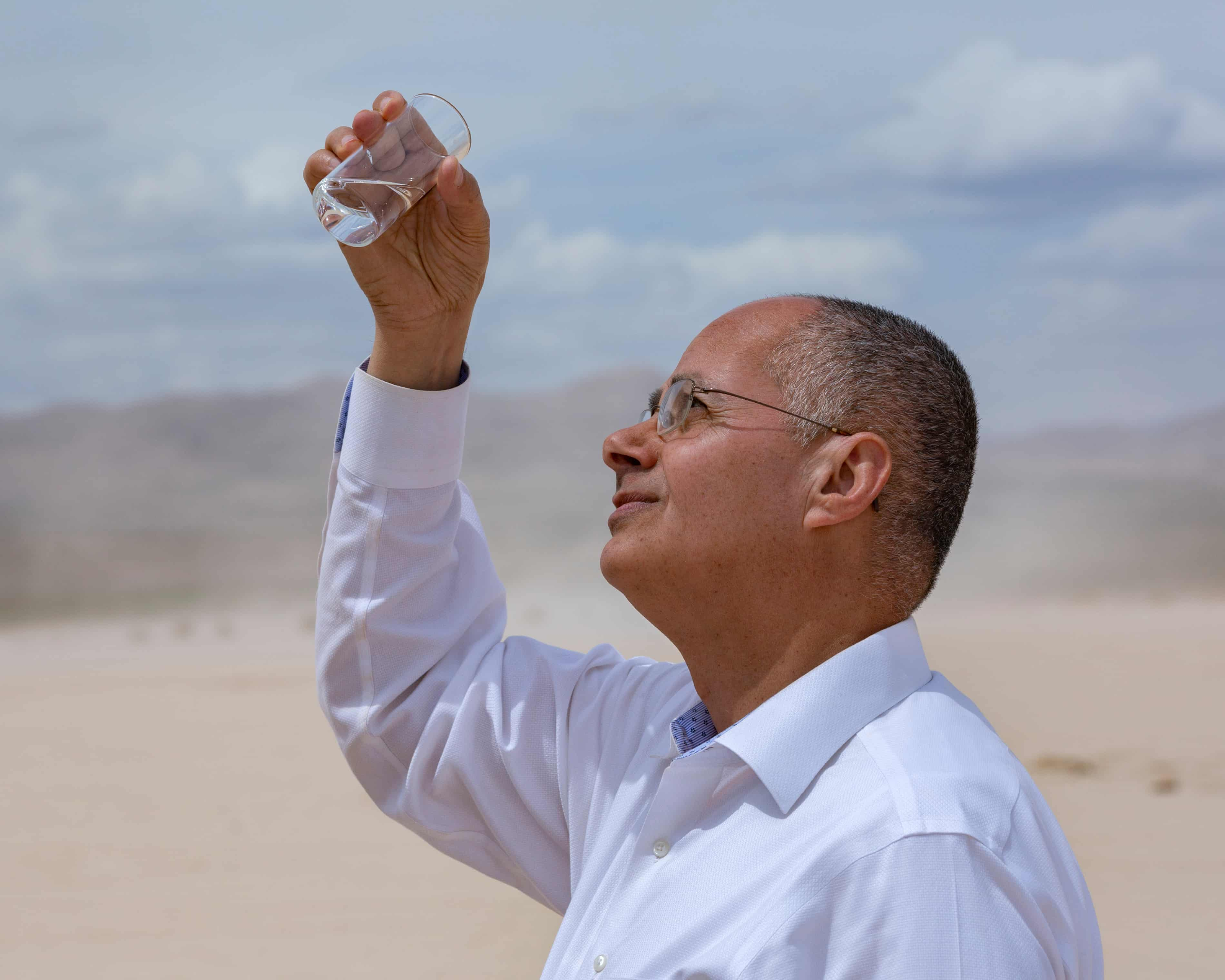 Un homme examine un verre d'eau dans un désert sous un ciel partiellement nuageux.