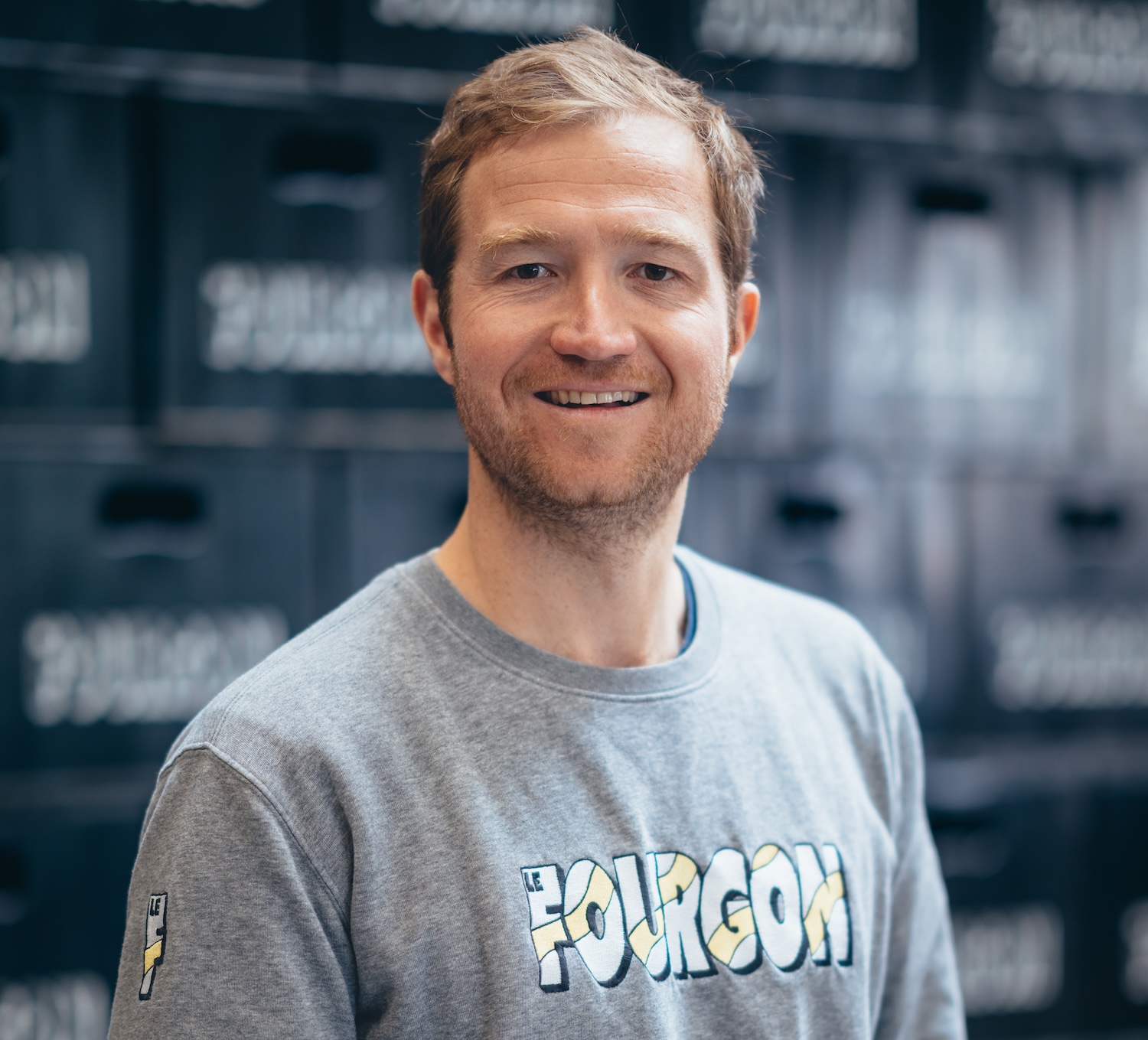 Homme souriant devant des caisses empilées, portant un pull gris, ambiance décontractée.