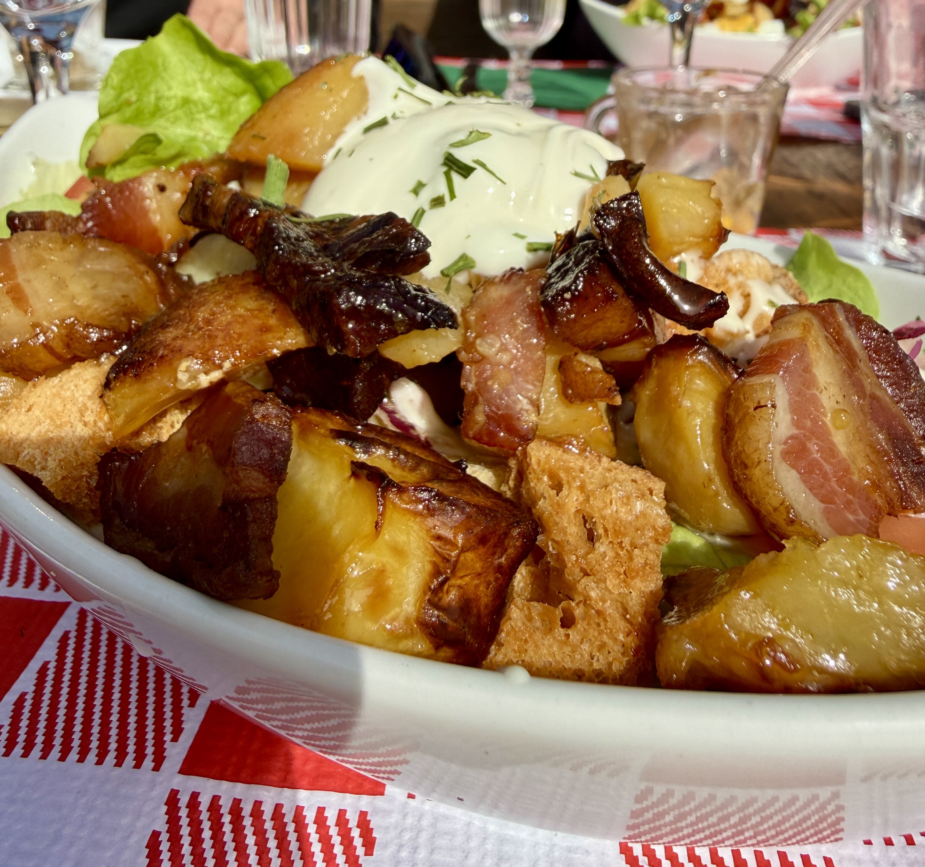 Plat de pommes de terre rôties, lardons, croûtons et salade, sur une nappe rouge.