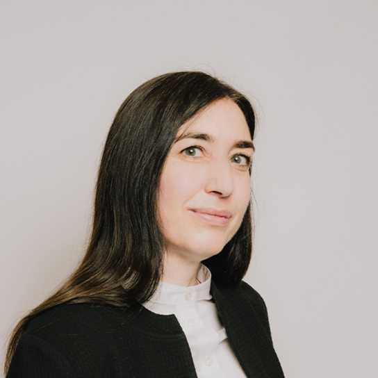 Femme aux cheveux noirs, souriante, portant une veste noire devant un fond clair.