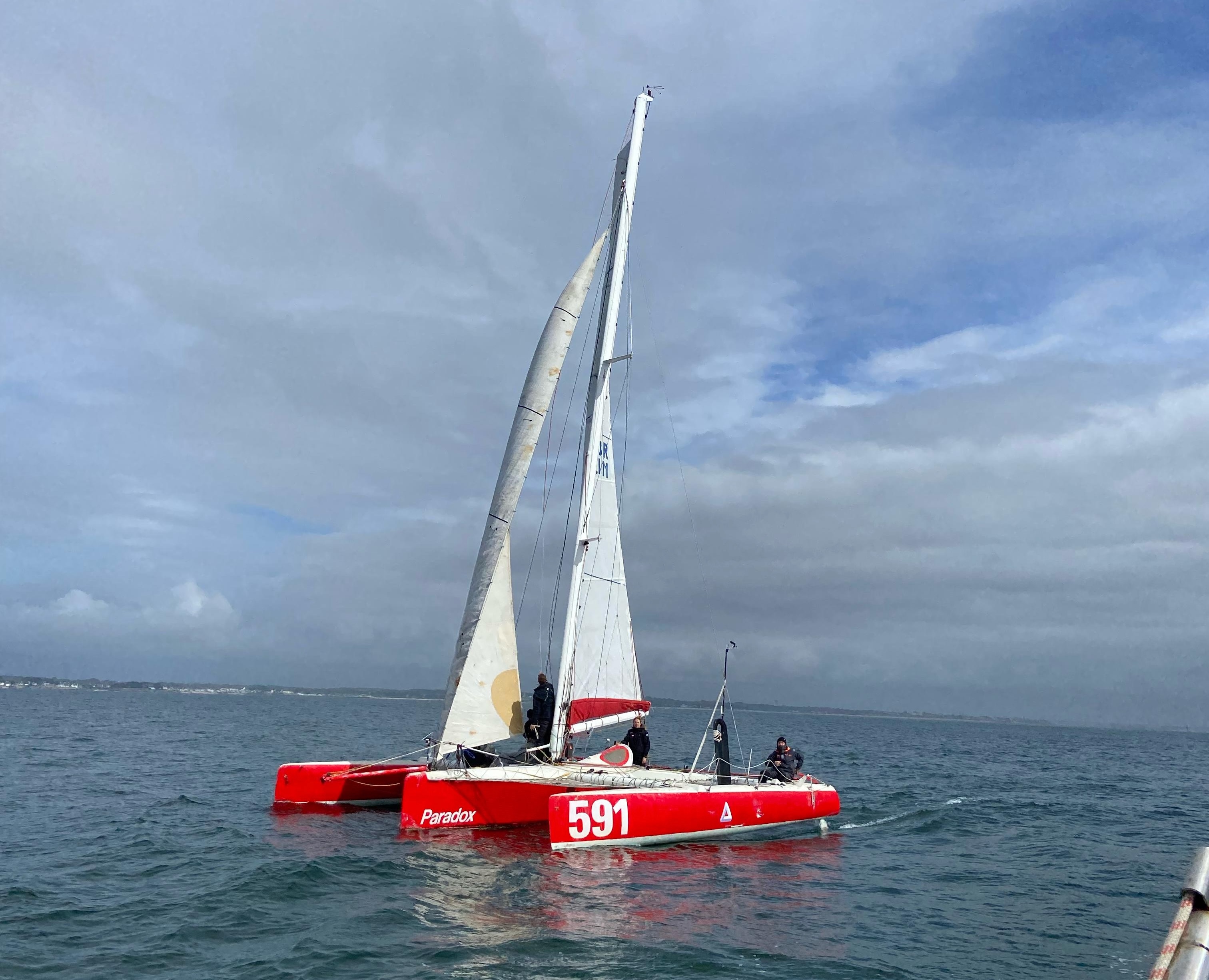 Voilier rouge sur l'eau avec équipage, ciel nuageux en arrière-plan.