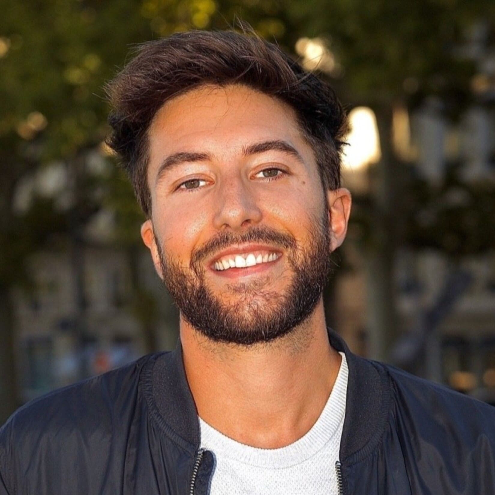Un homme souriant avec une barbe se tient dehors, éclairé par la lumière naturelle.