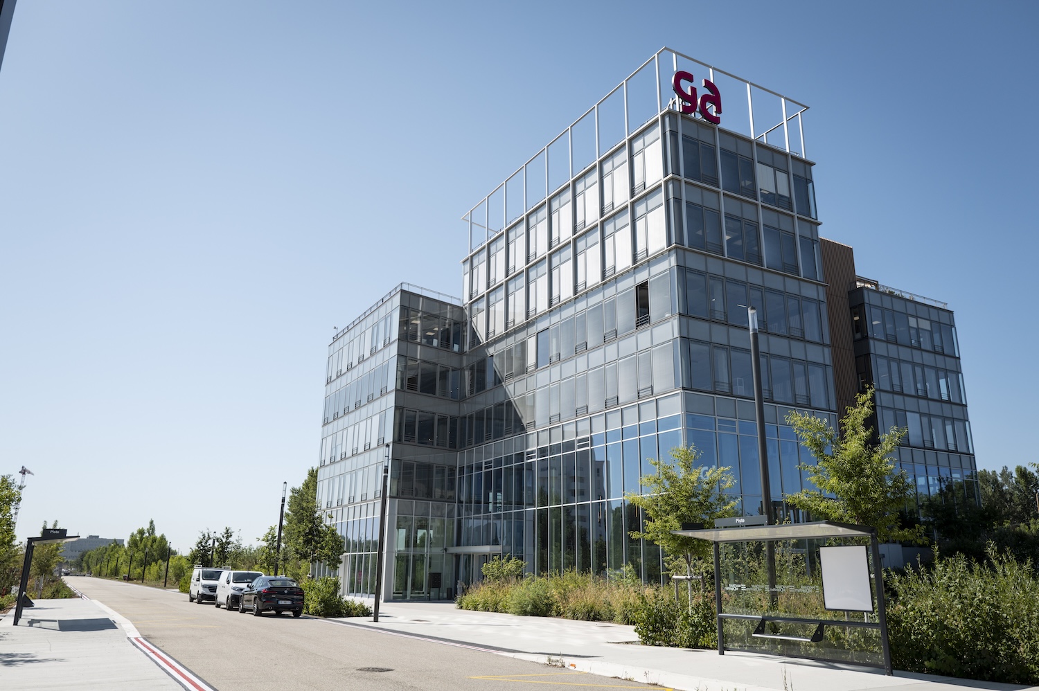 Bâtiment moderne en verre avec logo rouge sur le toit, arbres et voitures devant.