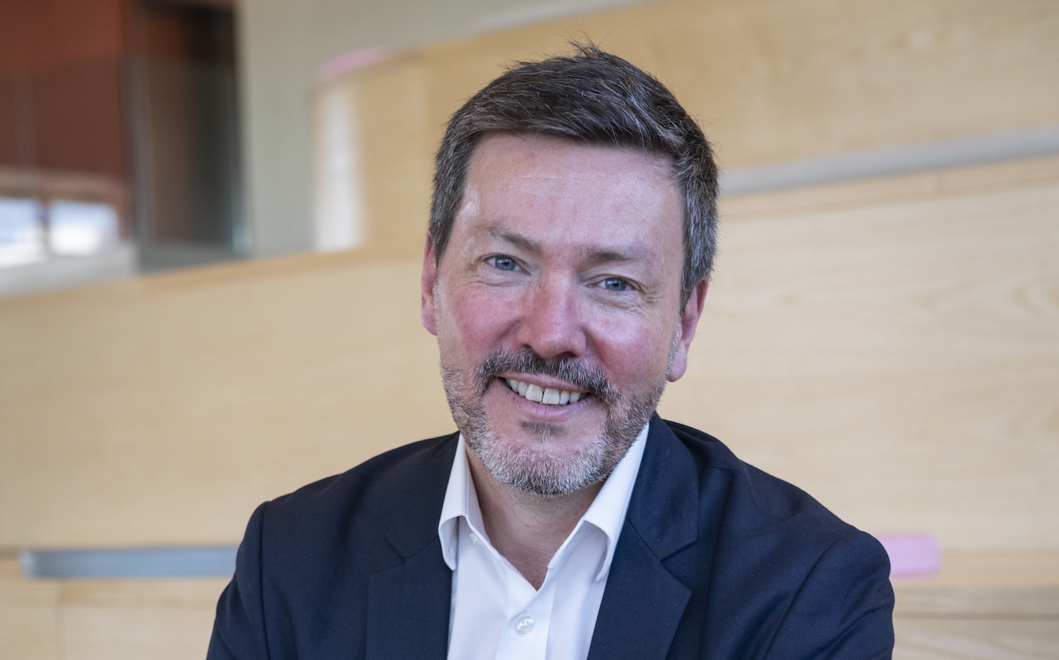 Homme souriant en costume assis devant un fond en bois.