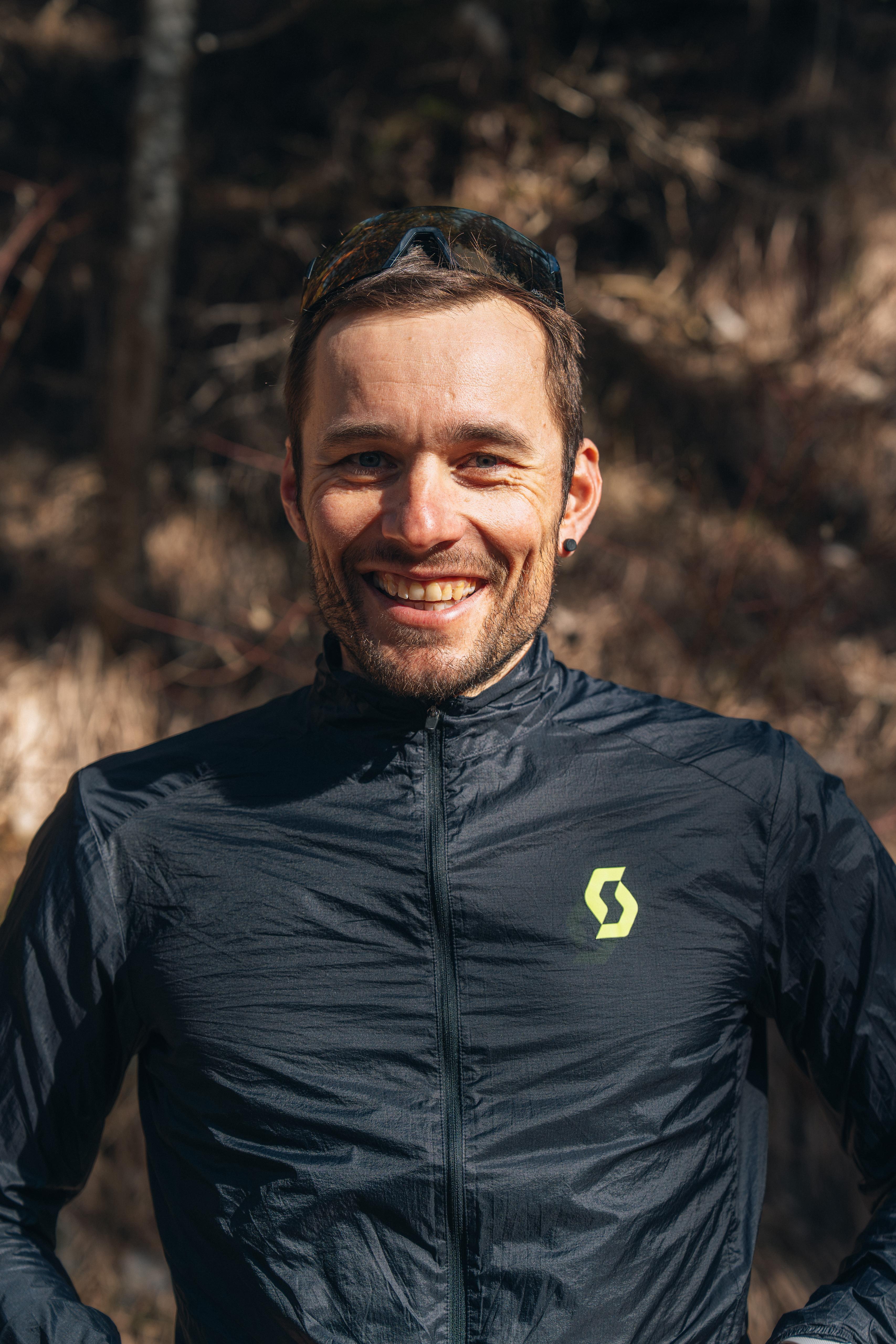 Homme souriant portant une veste noire en extérieur, avec un fond flou de nature.