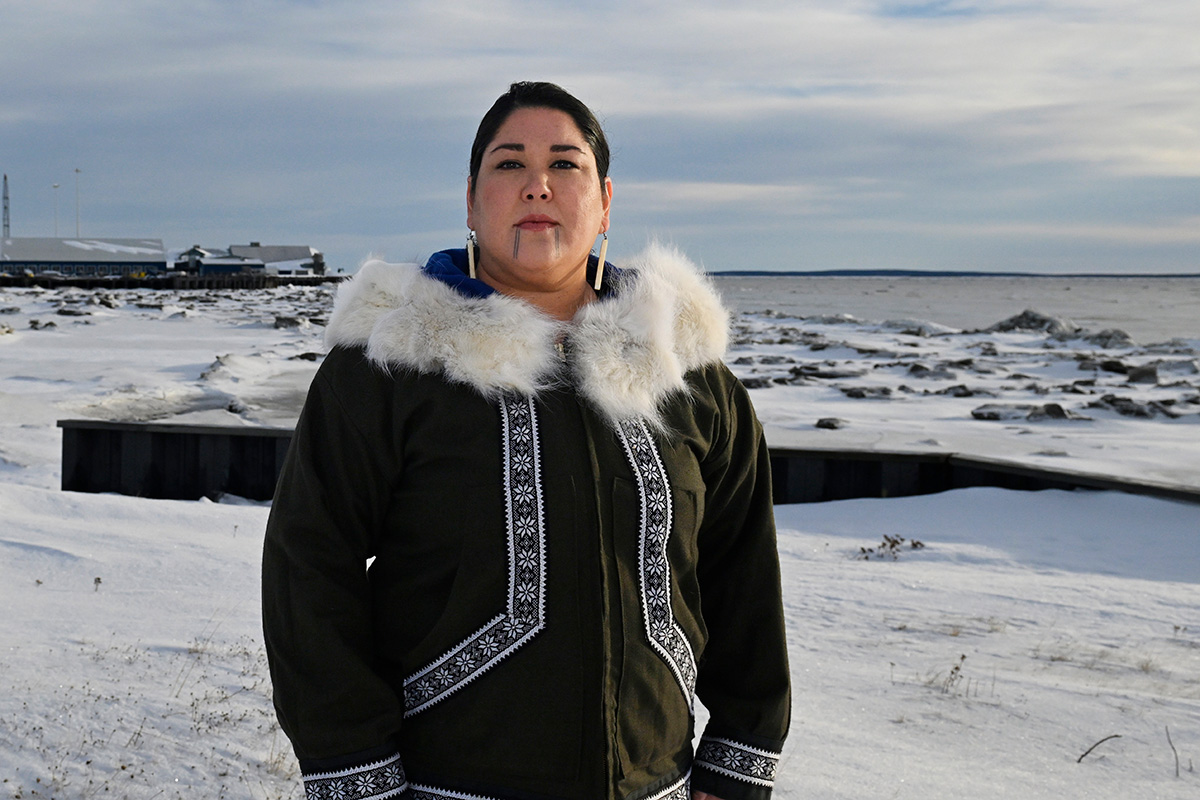 Personne en manteau traditionnel devant un paysage enneigé avec ciel nuageux et bâtiments éloignés.